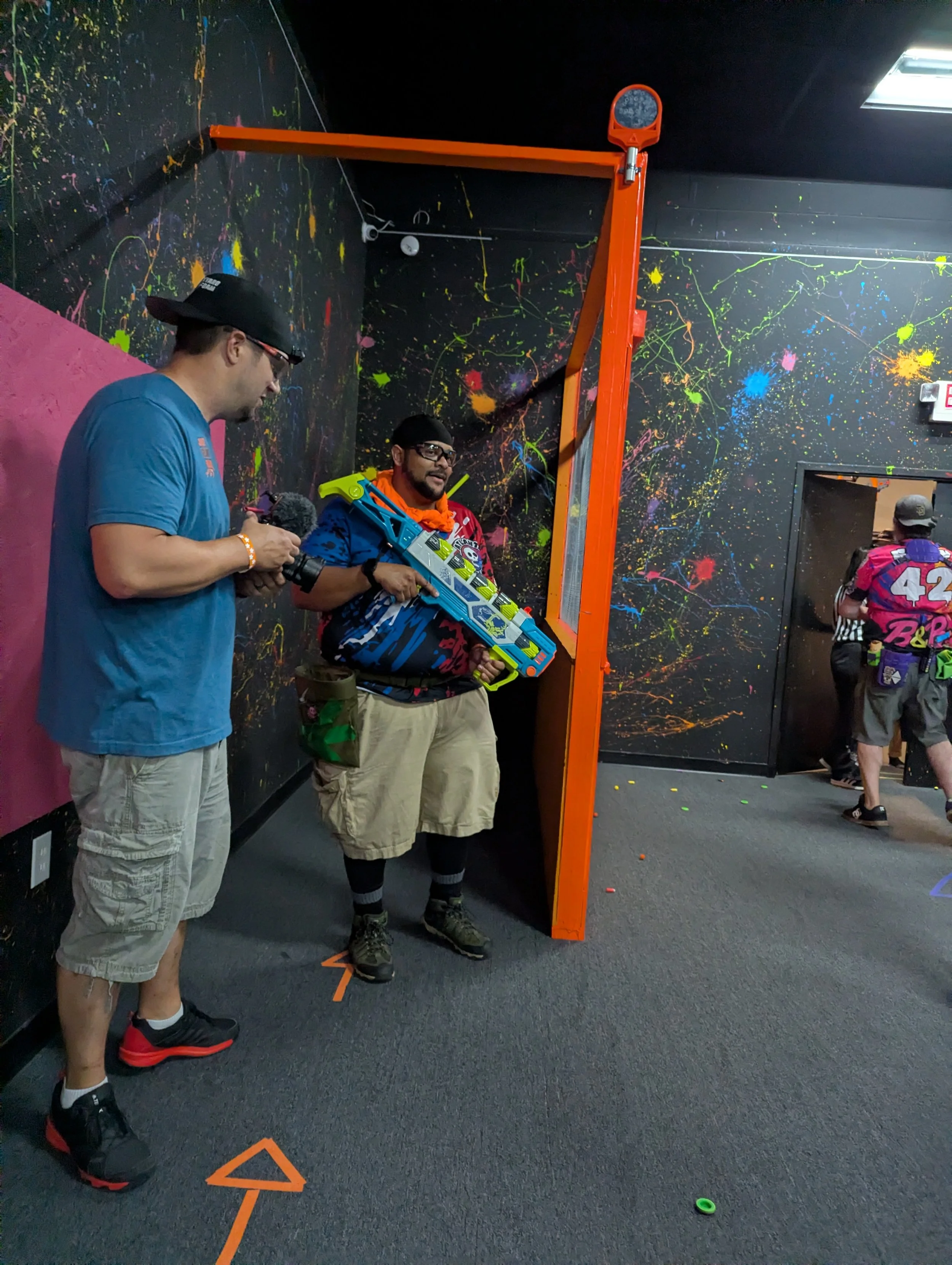 Two men playing an indoor activity using a foam nerf gun, one man is holding a nerf gun and the other man is standing with his hands in his pockets. The scene is decorated with colorful wall paint splashes, and a basketball hoop is mounted on the wal