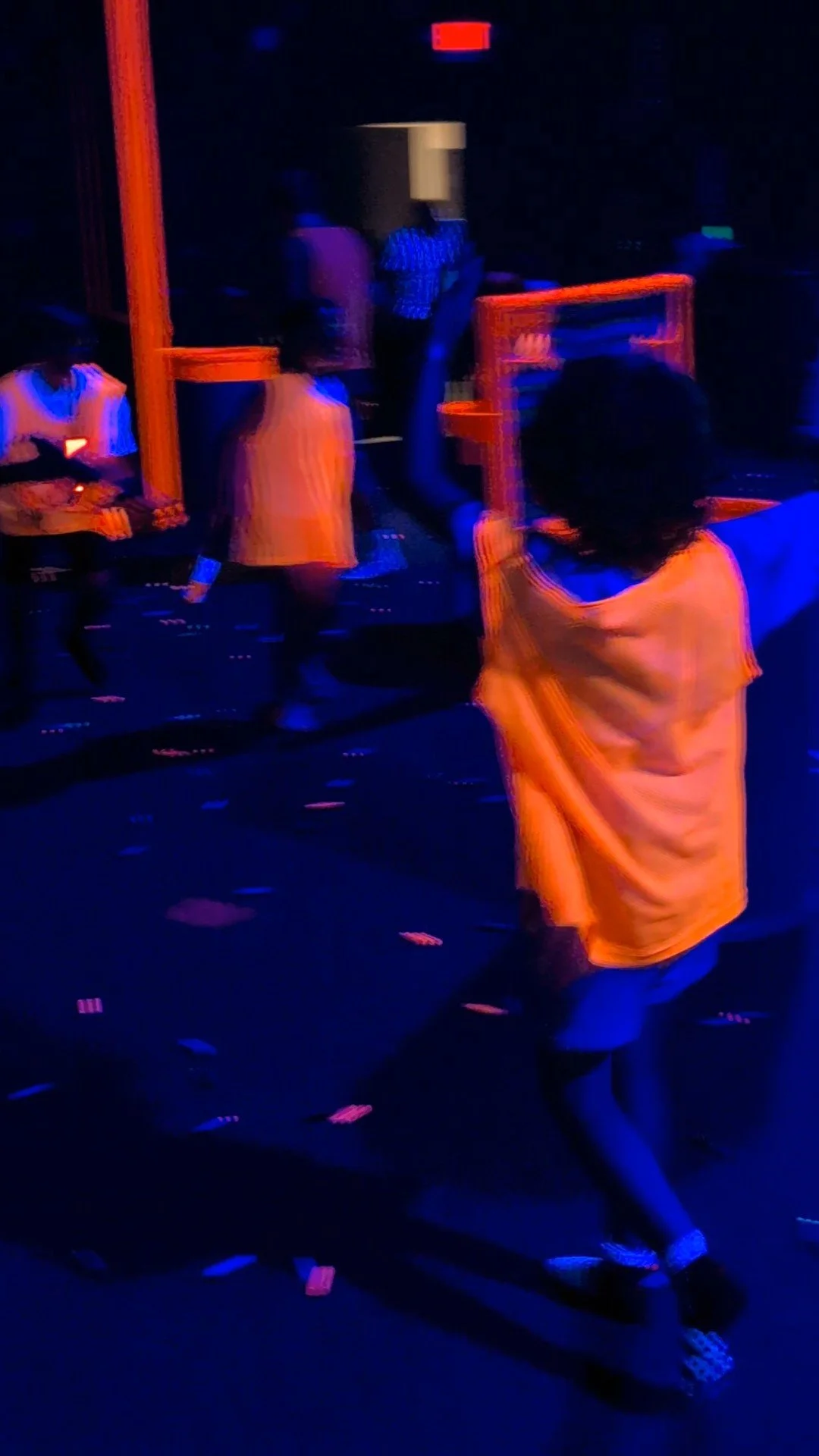 Children playing laser tag in a dark room with blue and orange lighting, some children are aiming lasers while others are running or hiding.