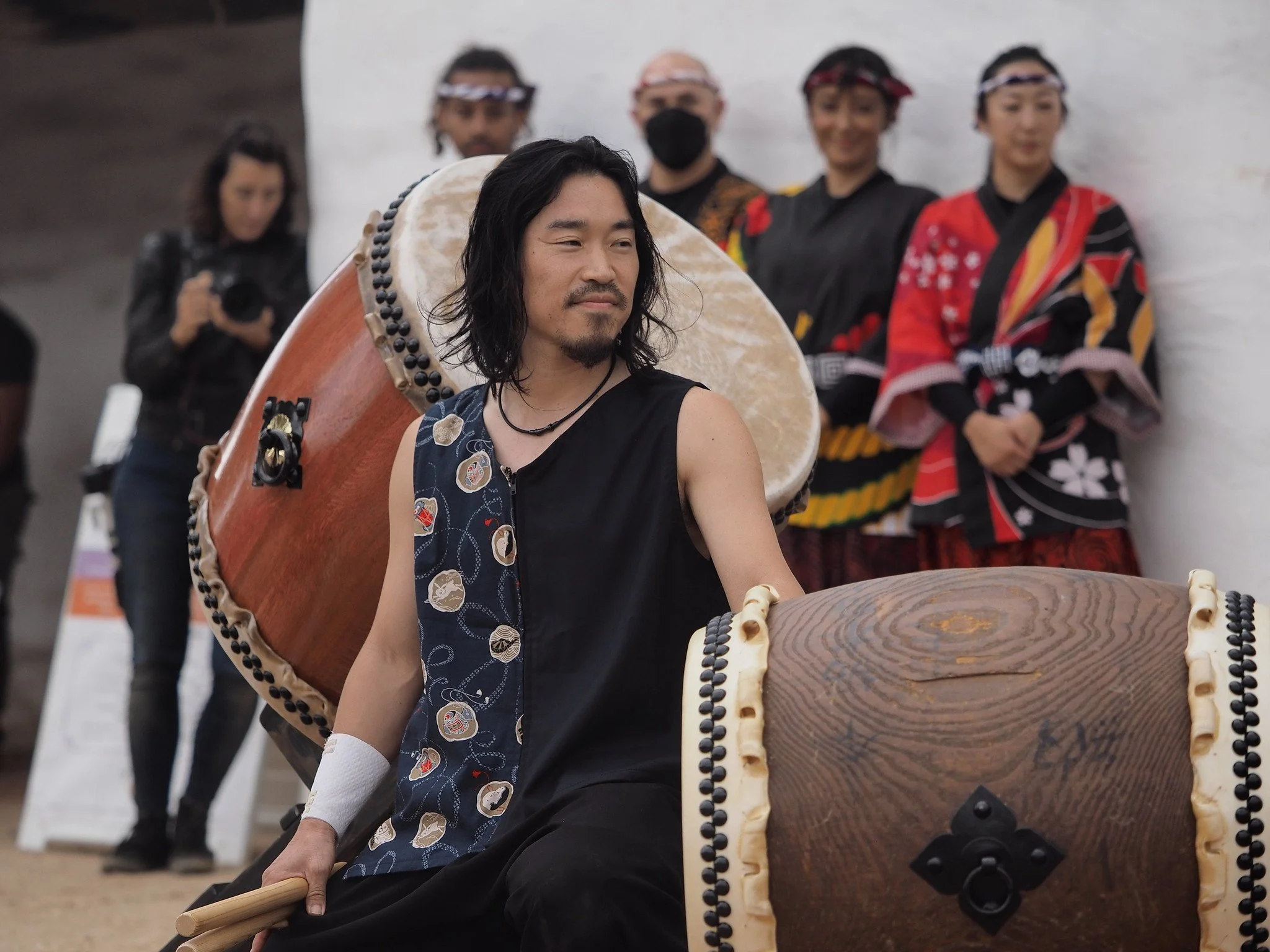 A man with long dark hair playing a large traditional drum, with three women dressed in colorful kimonos standing behind him, and a woman taking a photo in the background.
