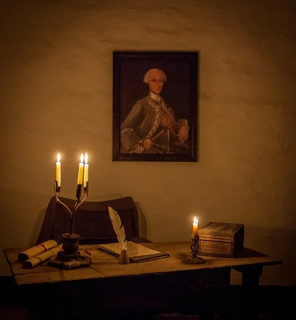 Vintage writing desk with candles, quill, open book, and portrait hanging above in dim lighting.