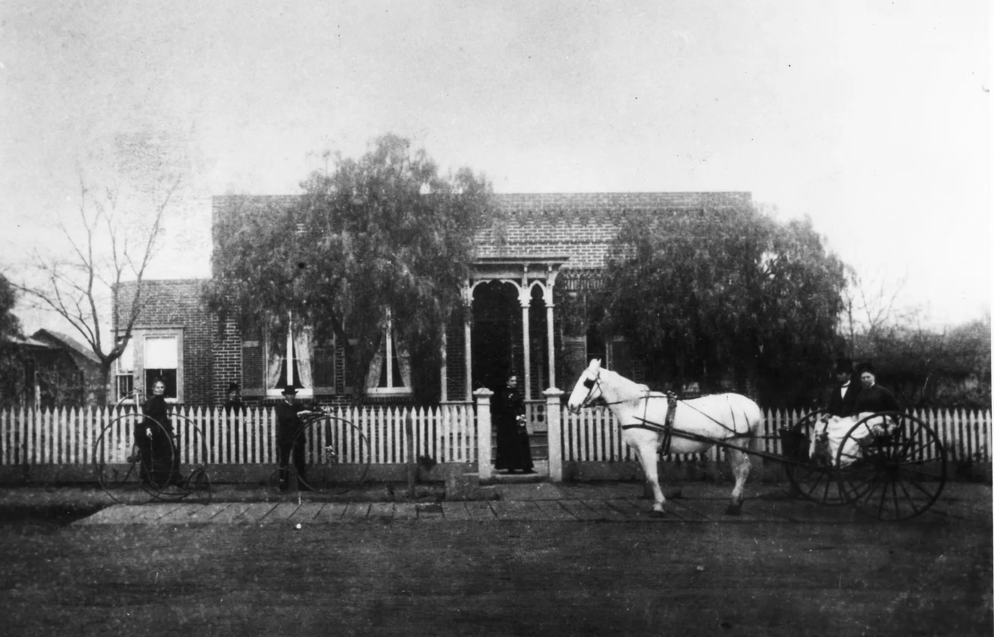 Black and white photo of a house with a porch, surrounded by a picket fence. Two women are standing beside a horse-drawn cart, and a man is sitting in the cart. The house has trees and leafless branches in the yard.
