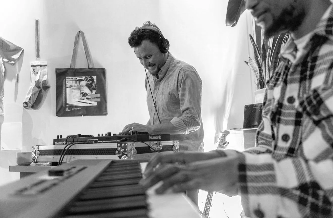DJ and musician performing together, one wearing headphones and operating a mixer, the other playing a keyboard, in a room with casual decor.