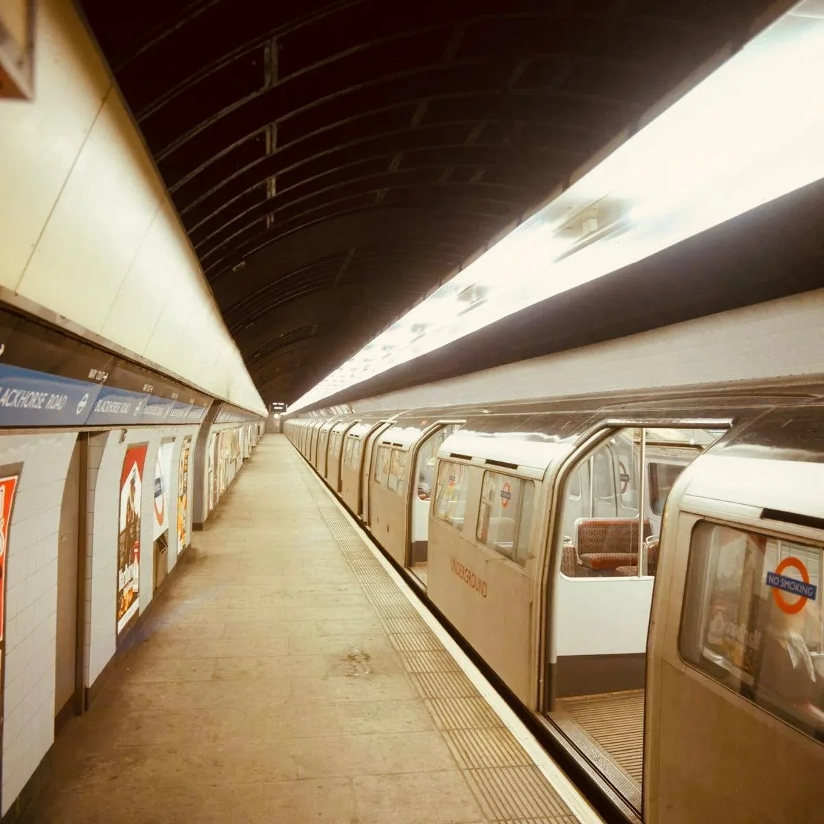 Underground subway platform with a train stopped at the station, featuring seating interiors visible through open doors, advertising posters, and 'Underground' signs.
