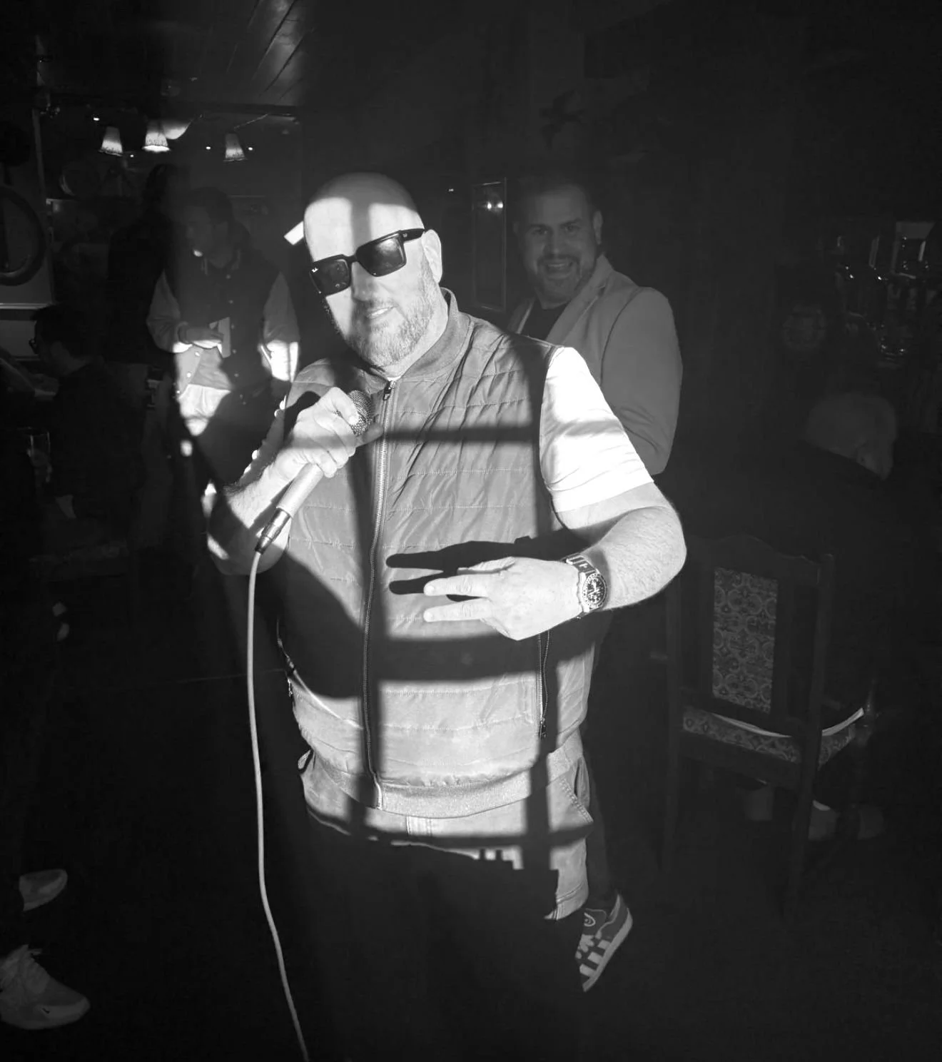 A man wearing sunglasses and a vest holds a microphone in a dimly lit room, while another person stands smiling in the background.