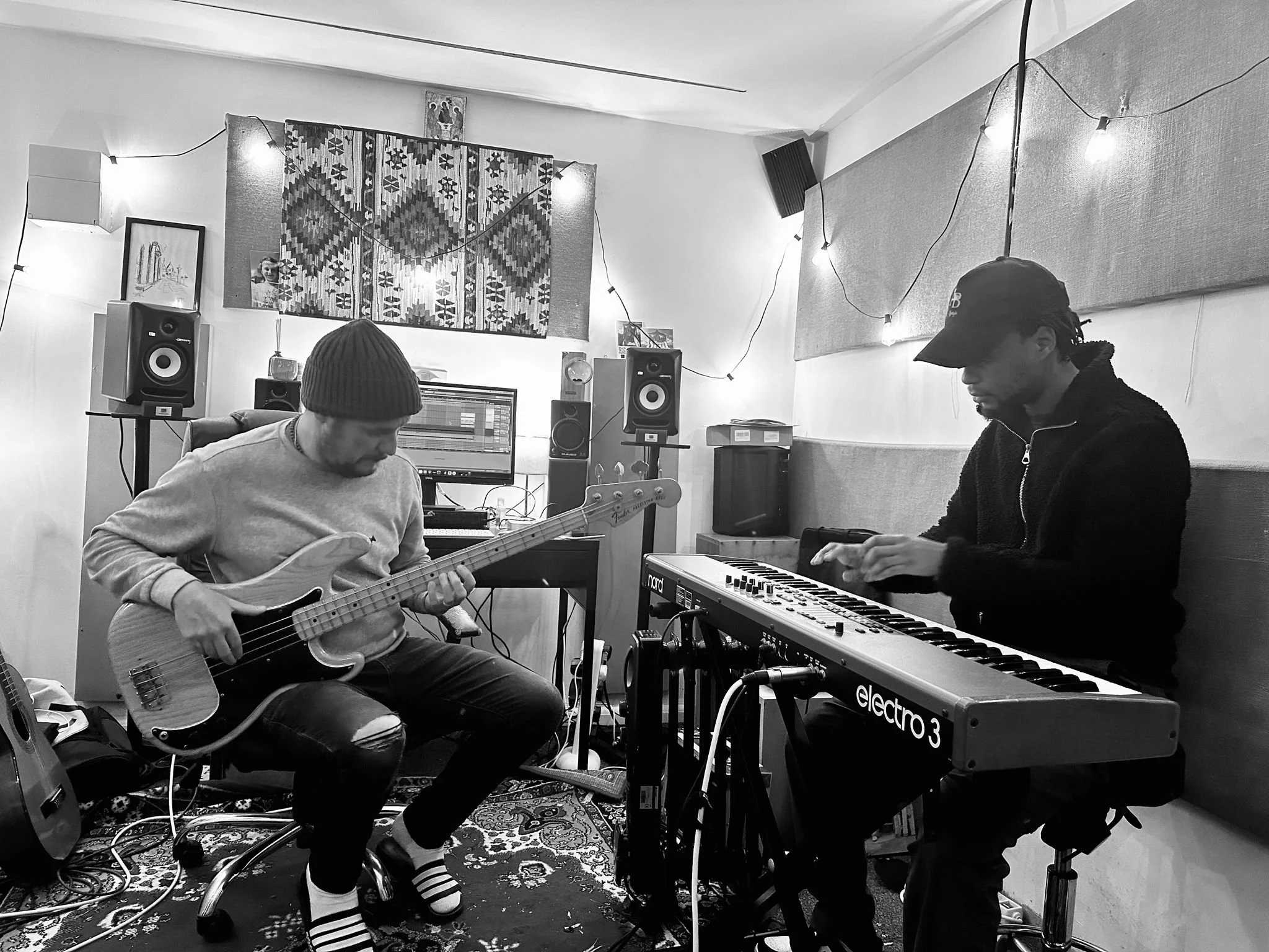 Two musicians in a recording studio; one playing an electric bass guitar and the other playing a keyboard while sitting on chairs. The room has studio monitors, a computer, and decorative lighting.