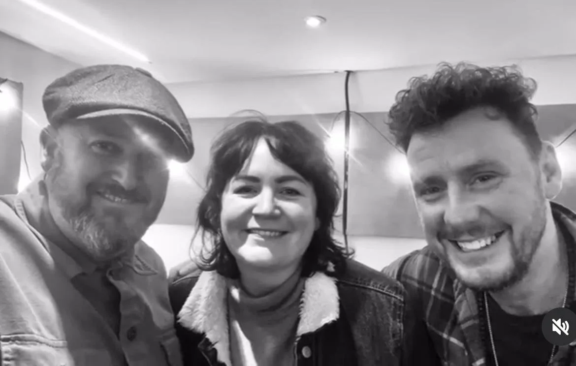 Three people smiling in a black and white photo, with one wearing a flat cap, one in the middle wearing a jacket, and the other in a checkered shirt. Background shows string lights.