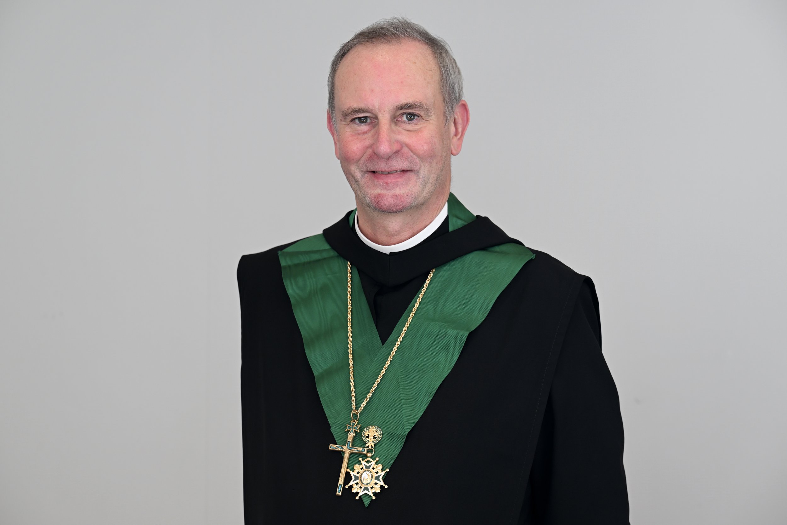 A man dressed in academic regalia with a green sash and multiple medals around his neck, standing against a plain gray background.