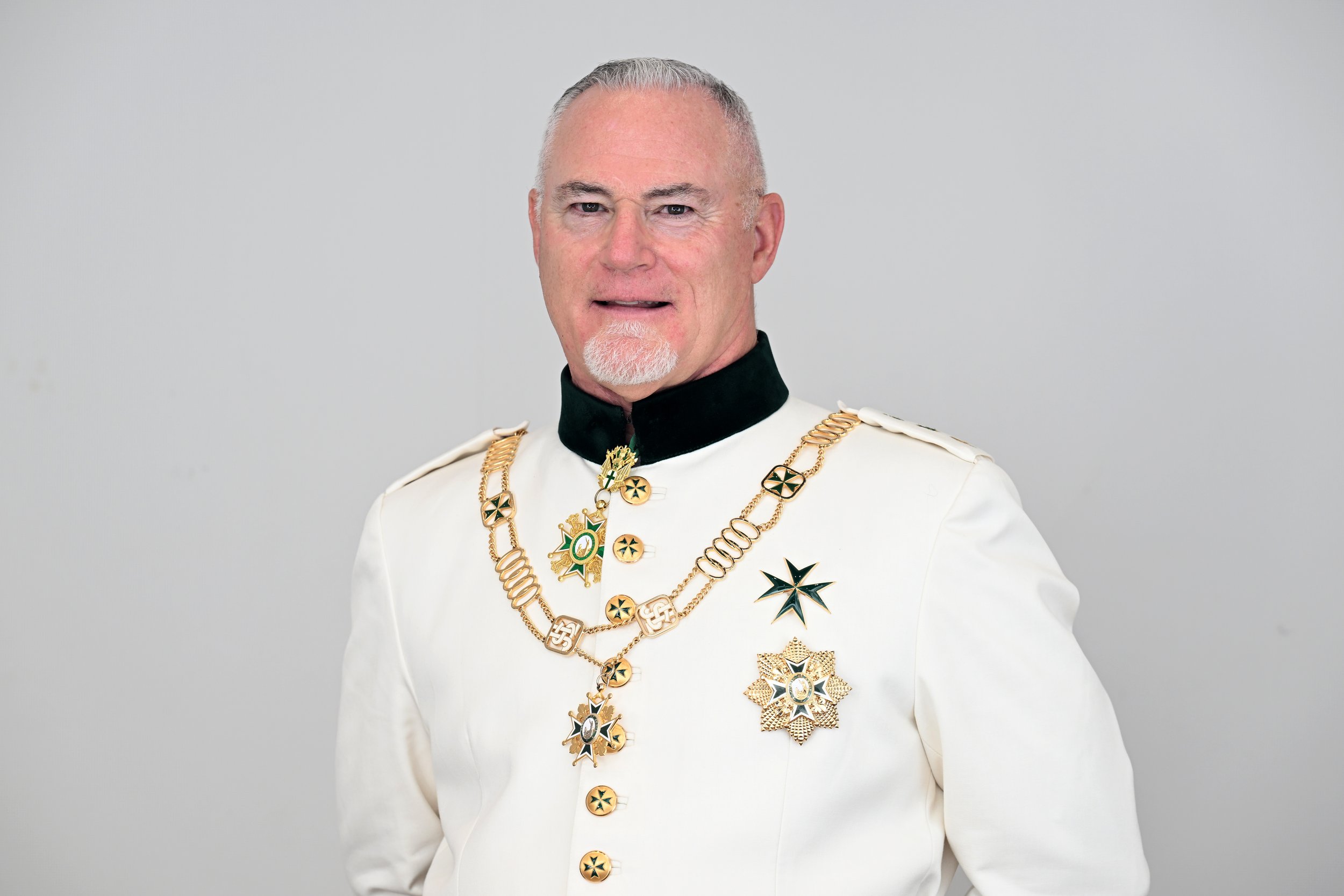 A man dressed in a white military-style uniform with a black collar, wearing multiple gold-colored medals and chains, standing against a plain light gray background.
