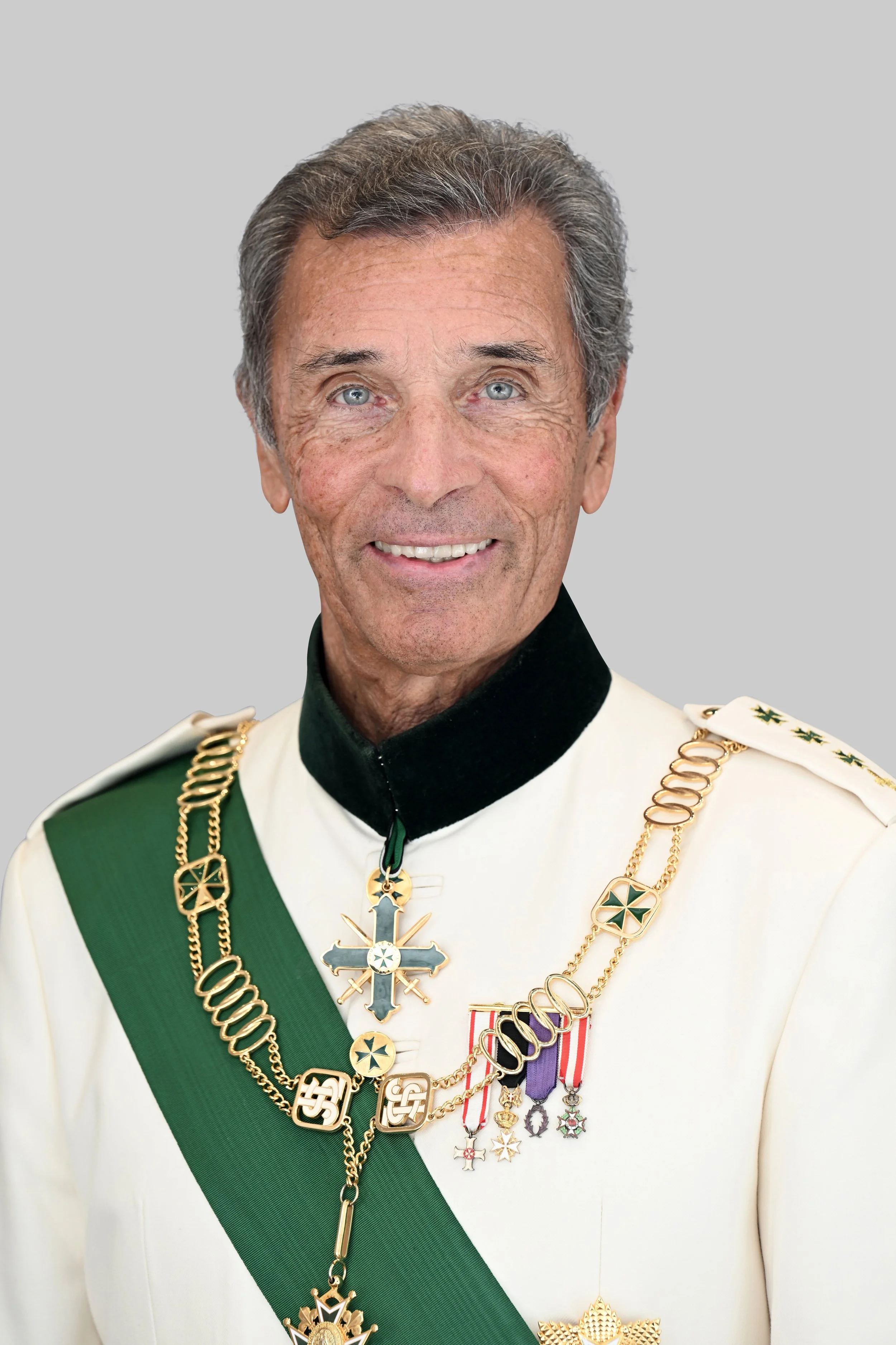 A man in formal military attire with a white uniform, black collar, green sash, and many medals and gold chains, smiling against a plain gray background.