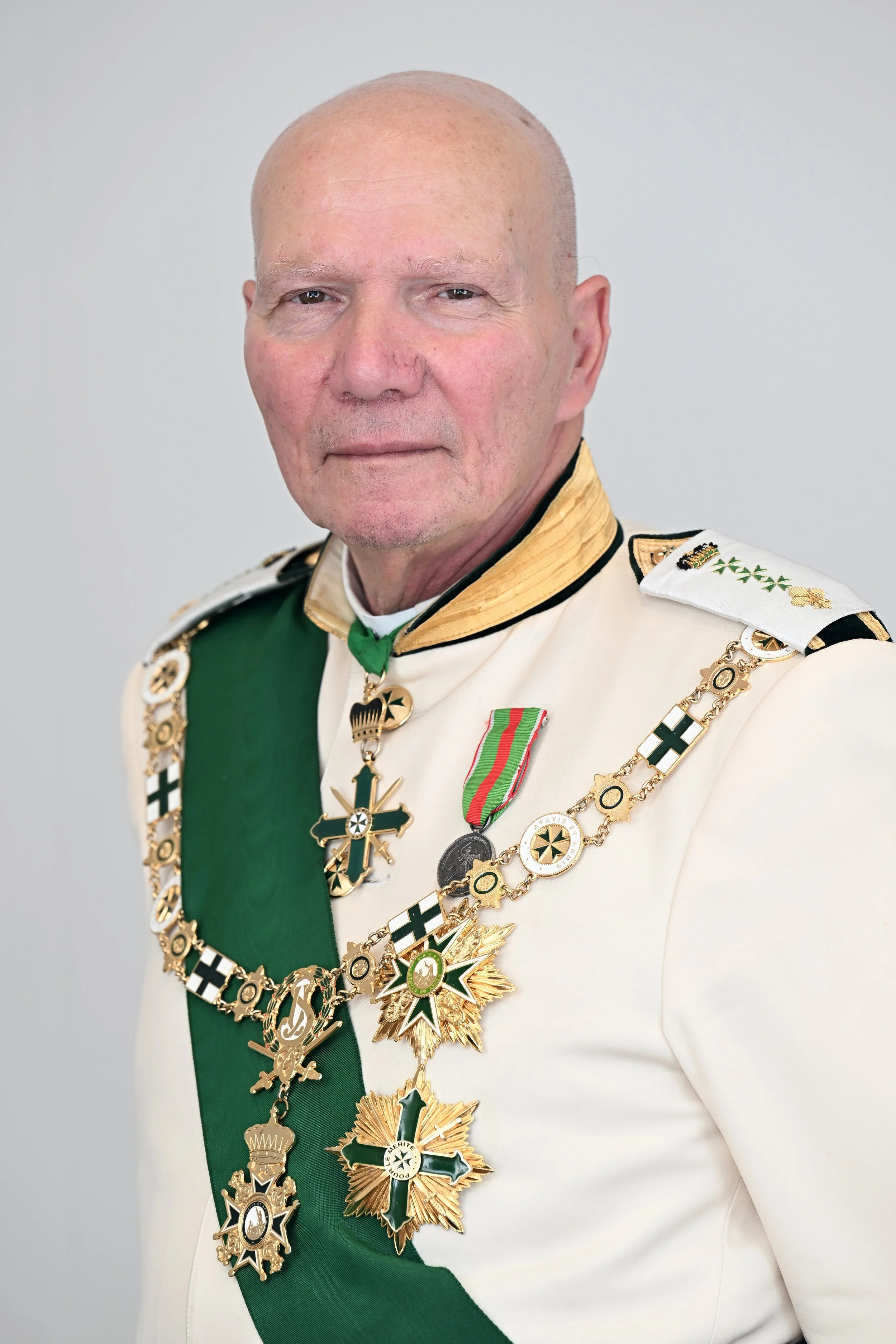 Elderly man wearing a white military uniform adorned with numerous medals and decorations, including a green sash, gold chains, and various crosses.
