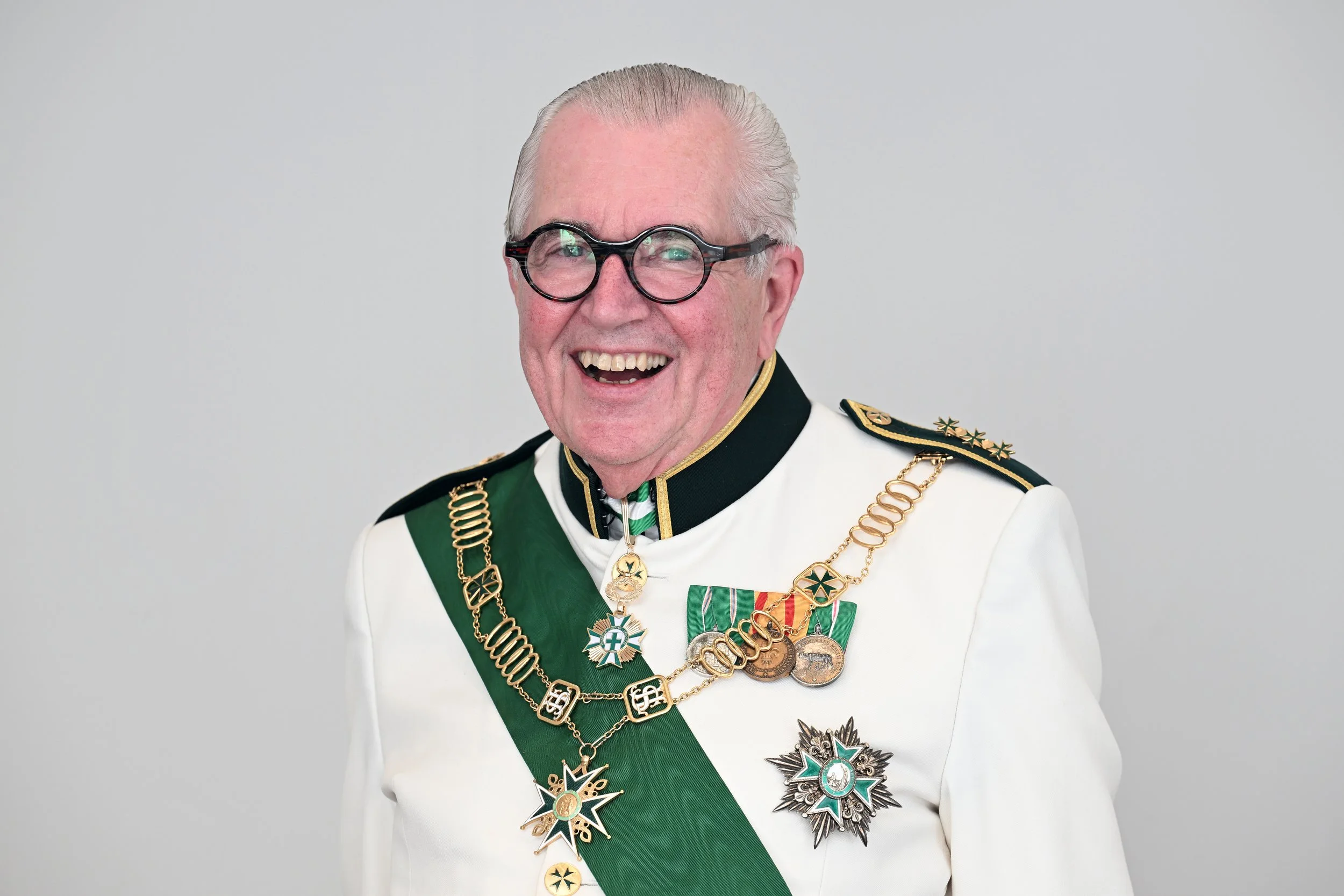 An elderly man in a military dress uniform adorned with medals, chains, and a green sash, smiling and wearing black-rimmed glasses.