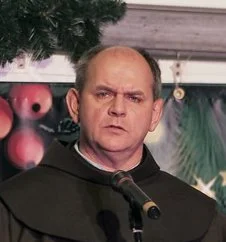 A man with a serious expression stands behind a microphone, with Christmas decorations in the background.