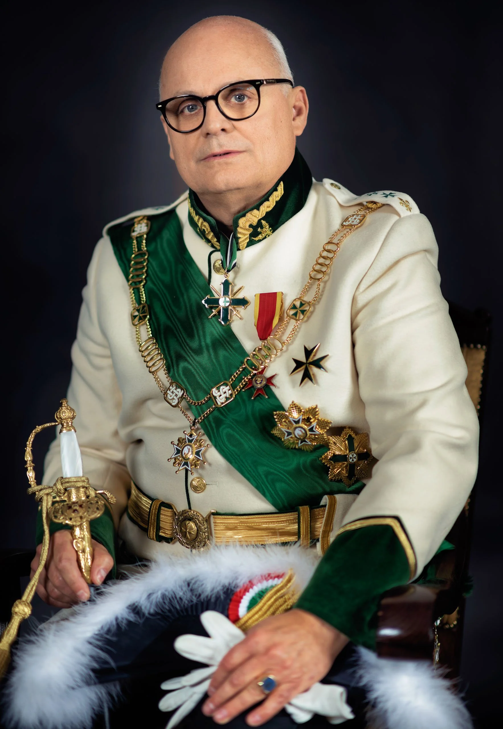 A man dressed in white military uniform decorated with medals and regalia, holding a sword, sitting in a chair against a dark background.