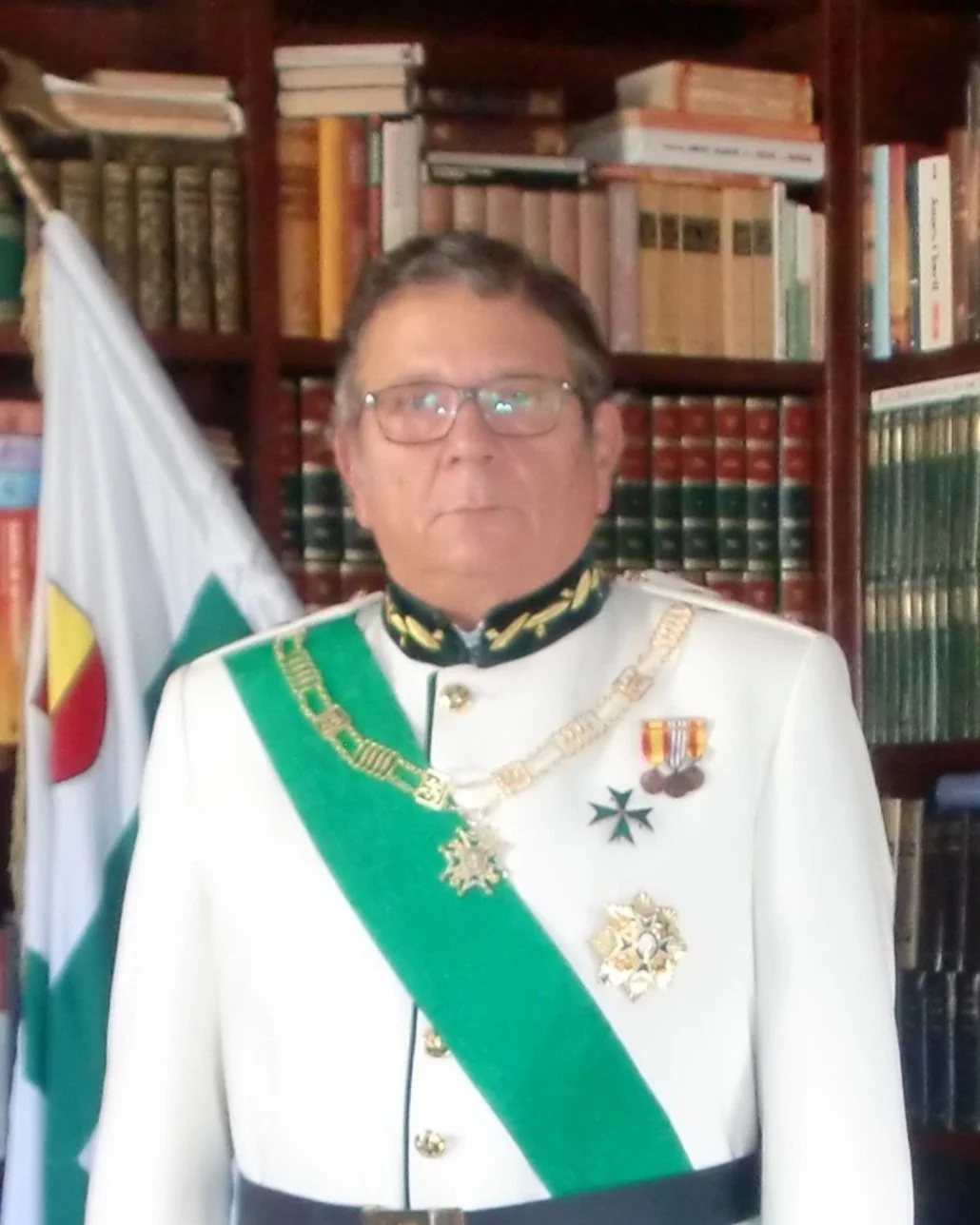 A man in a white military uniform with medals, wearing a green sash and chains, standing in front of bookshelves filled with books and a flag.