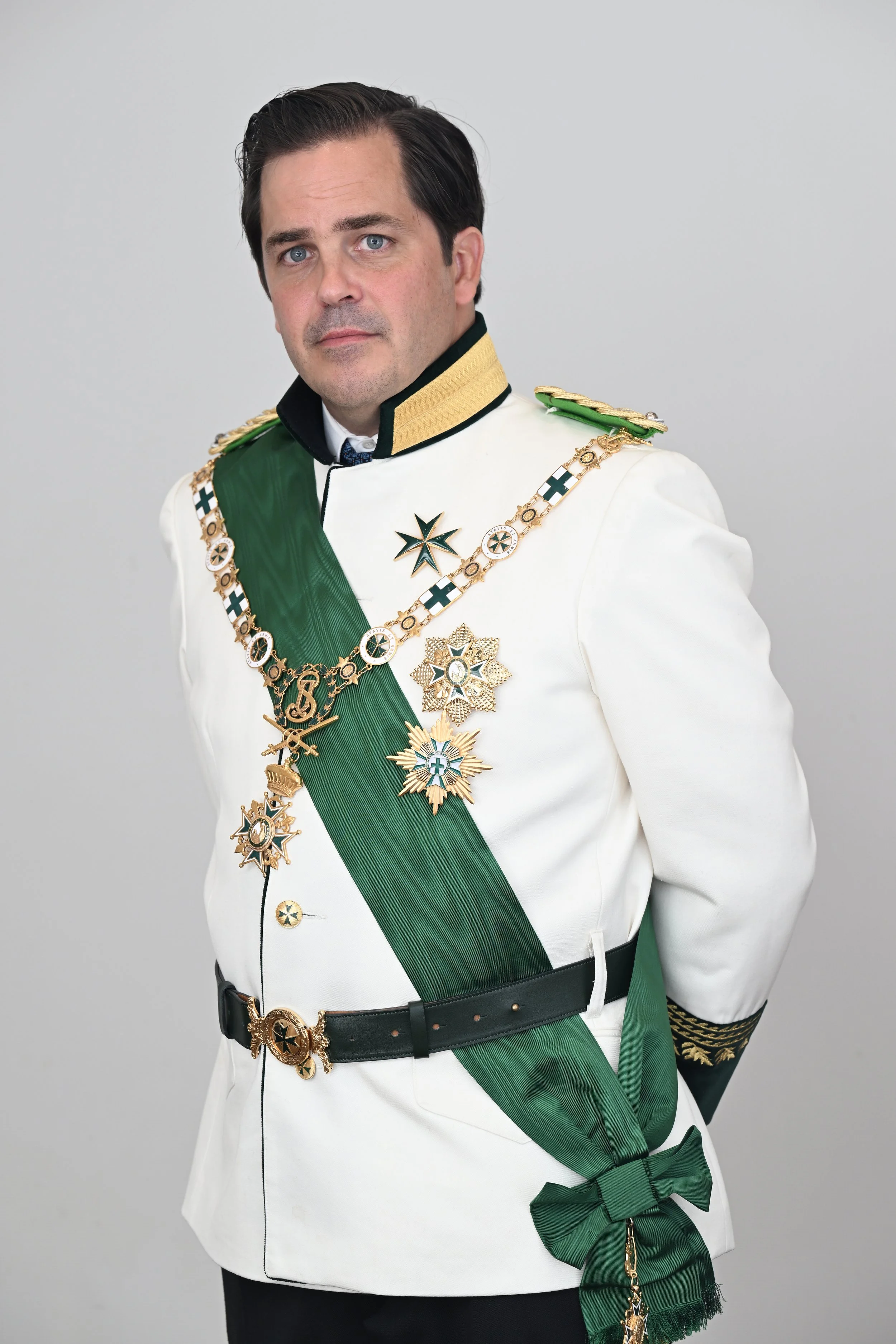 Man dressed in a military or formal official uniform with medals and a green sash, standing against a plain gray background.
