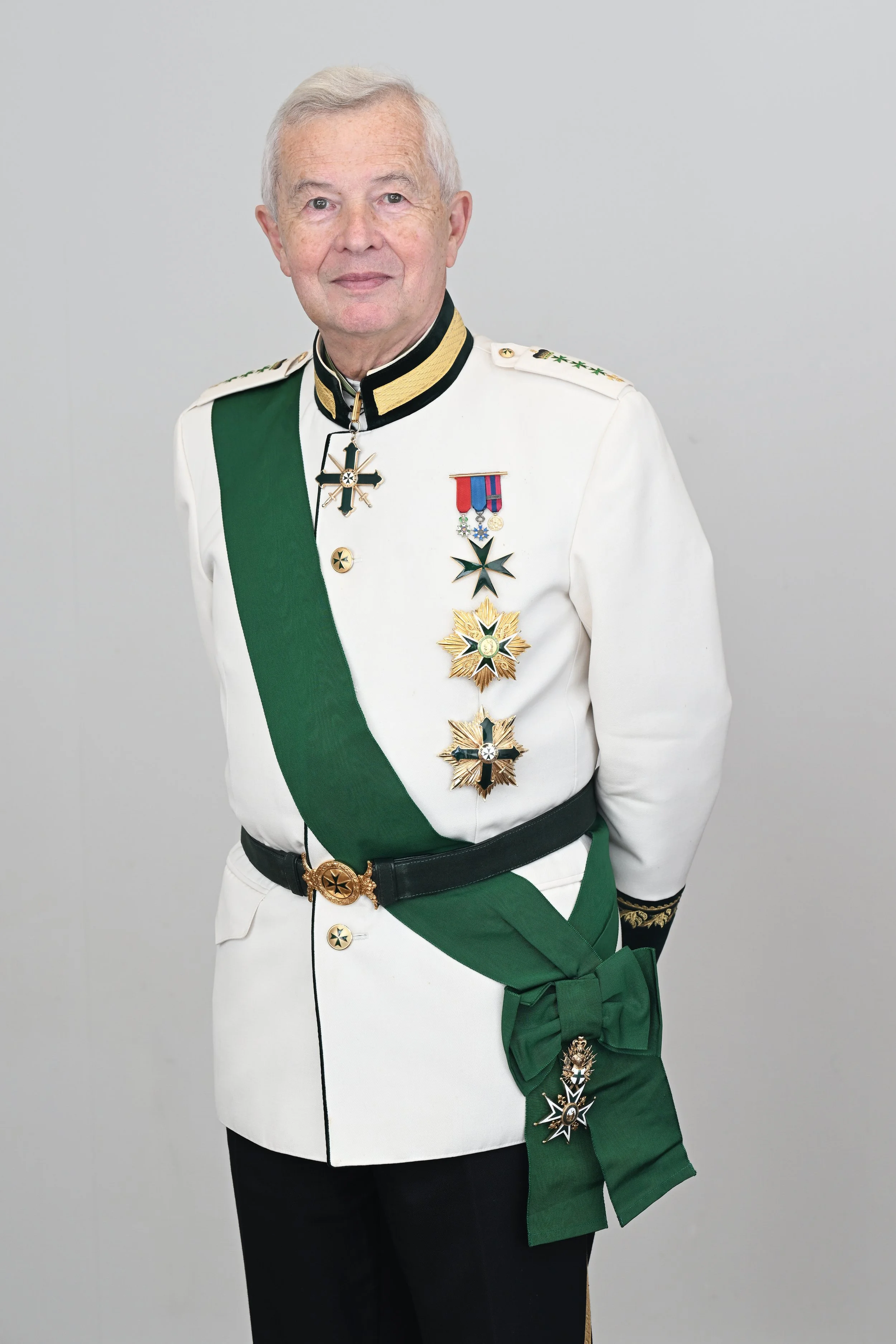 An elderly man in a white military uniform decorated with medals, a green sash, and a large green bow, standing against a plain light gray background.