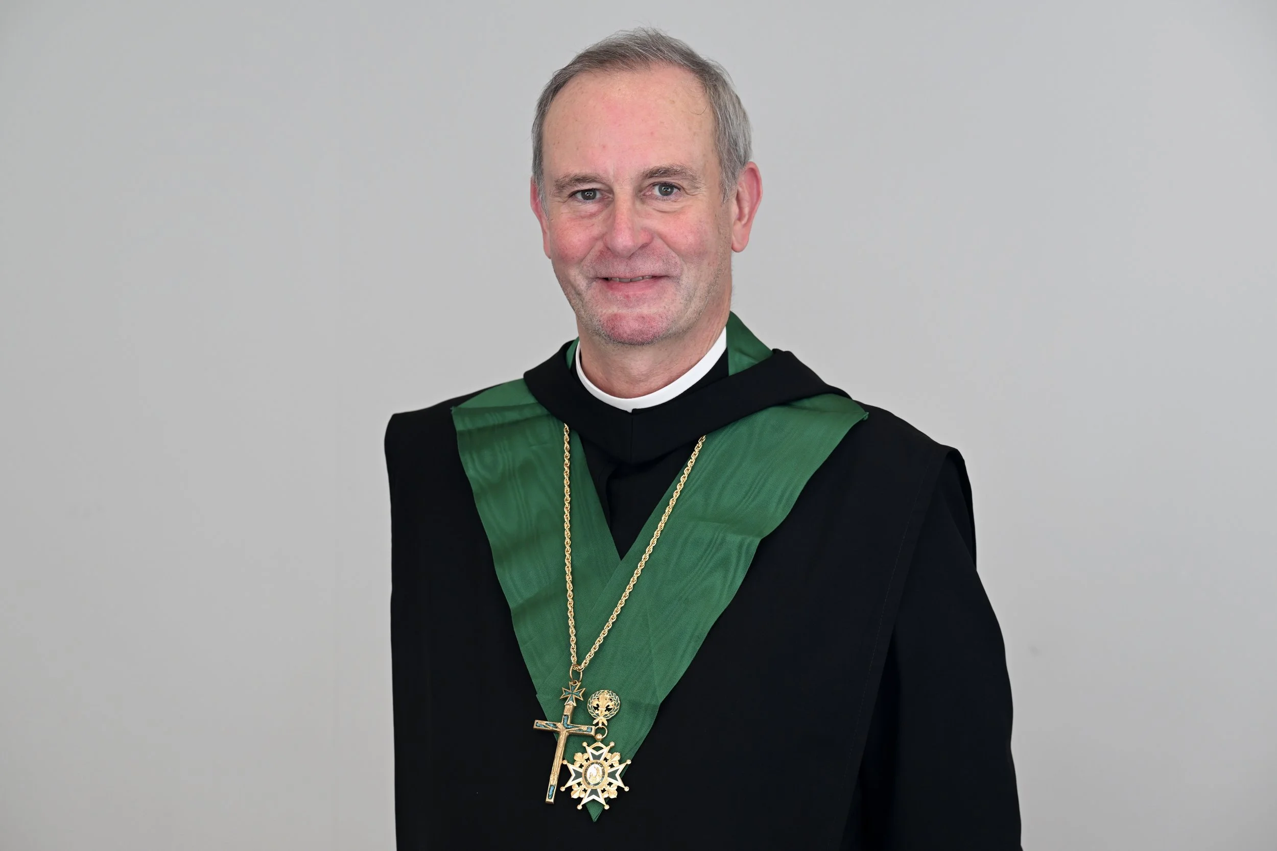 A man in academic regalia with a green and black stole, wearing multiple gold medals and chains, standing against a plain gray background.