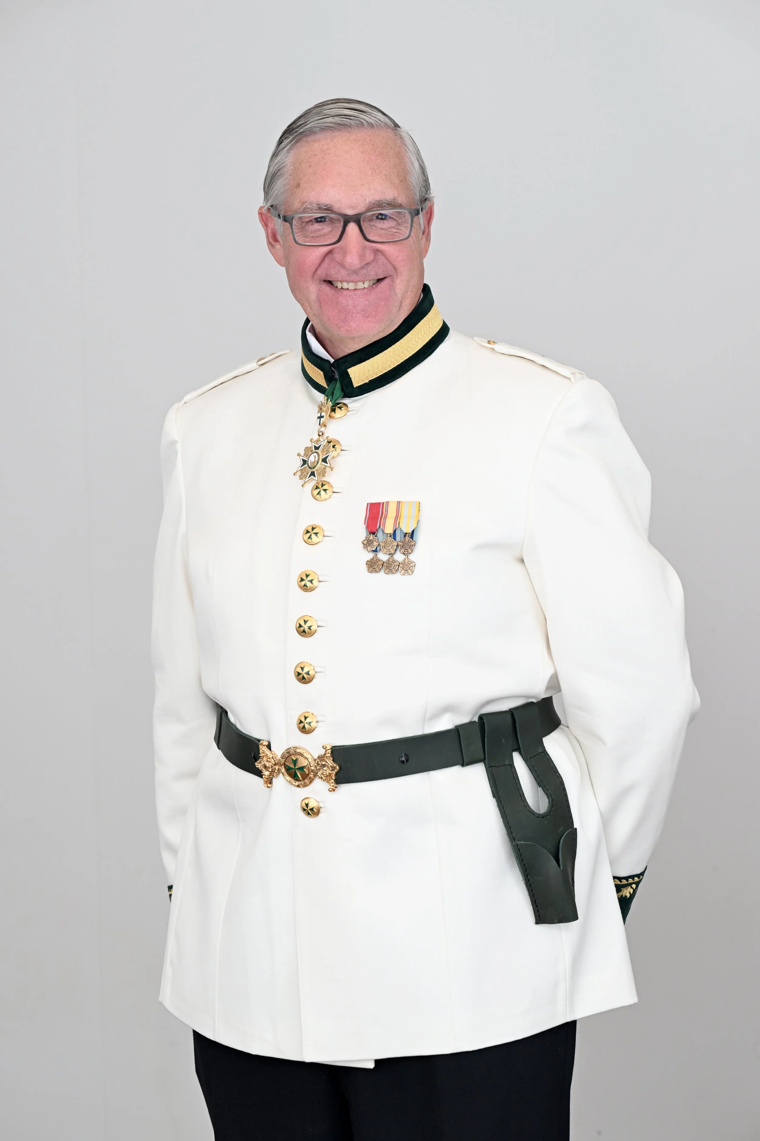 A smiling man in a white military uniform with medals and decorations, wearing glasses and standing against a plain gray background.