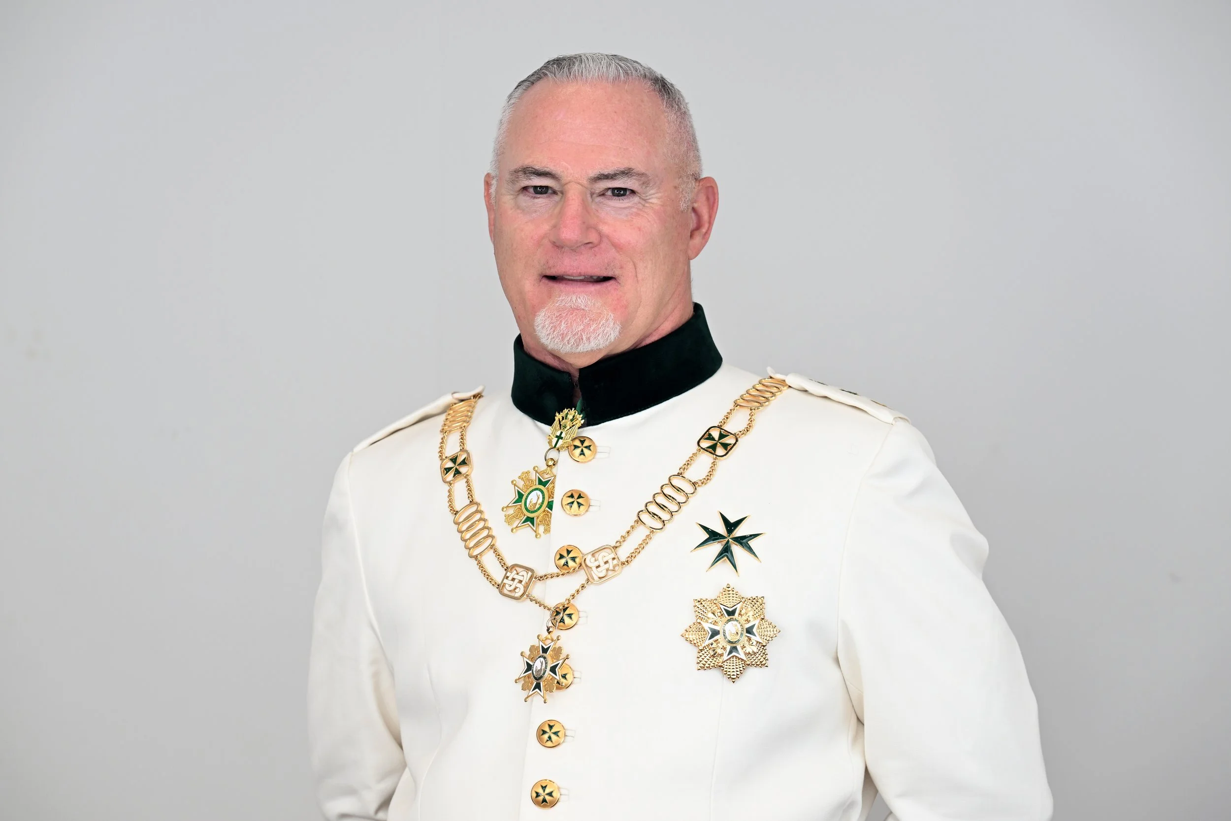 A man dressed in a white military-style uniform with black collar, adorned with multiple gold medals and decorations, standing against a plain gray background.