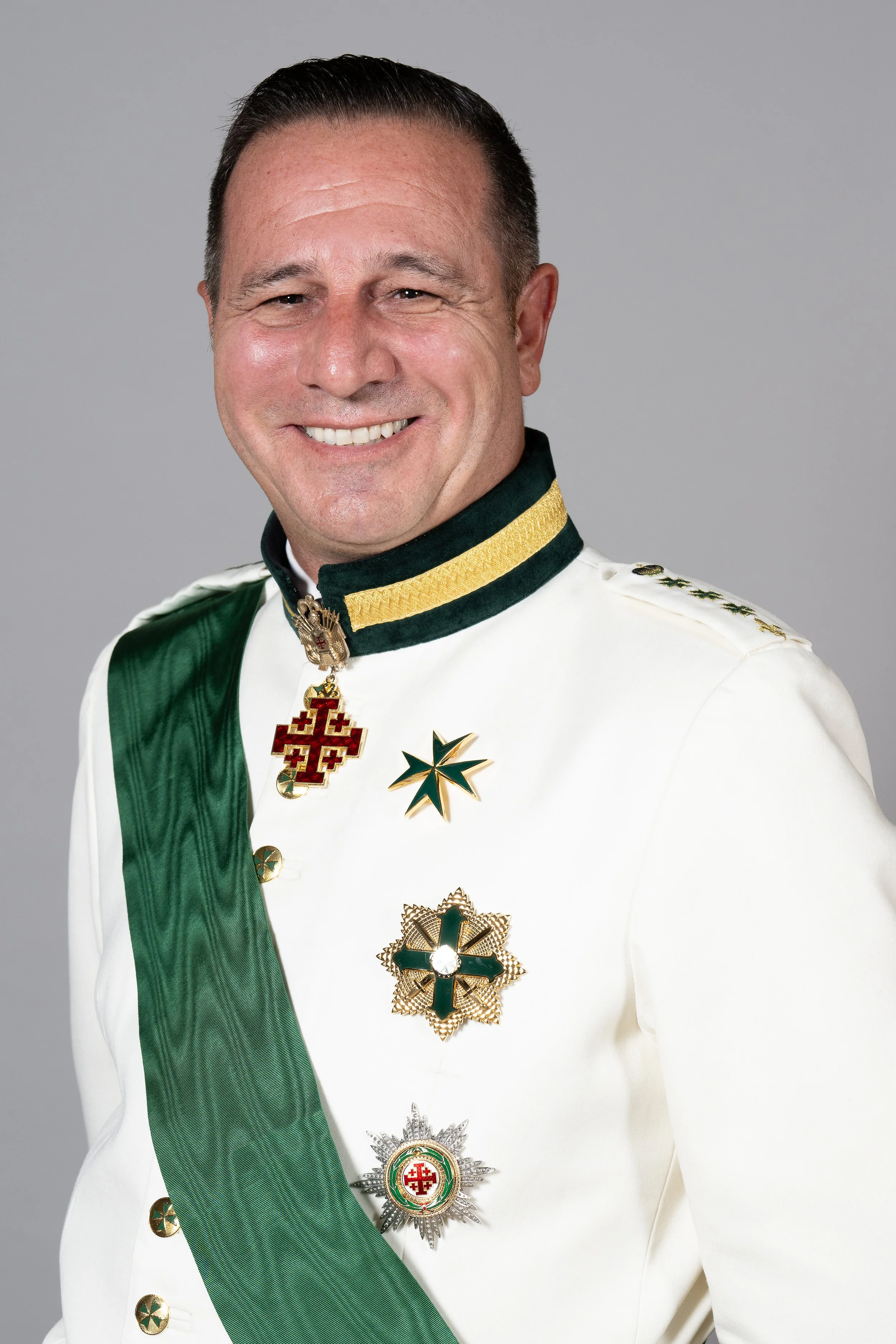 A smiling man wearing a white military uniform adorned with various medals and decorations, including a green sash, a green and yellow collar, and medals on his chest.
