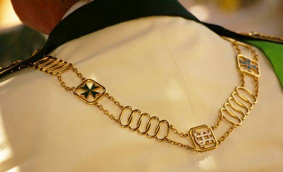 Close-up of a person's shoulder wearing a gold chain necklace with various charms, including an eight-pointed star, stacked loops, and a shield with intricate designs.