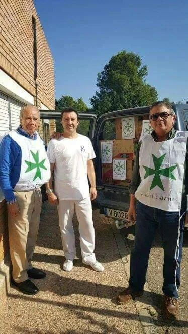 Three men standing outside next to a vehicle with boxes, wearing volunteer aprons with a green cross and the words 'Sant Lazare' on them.