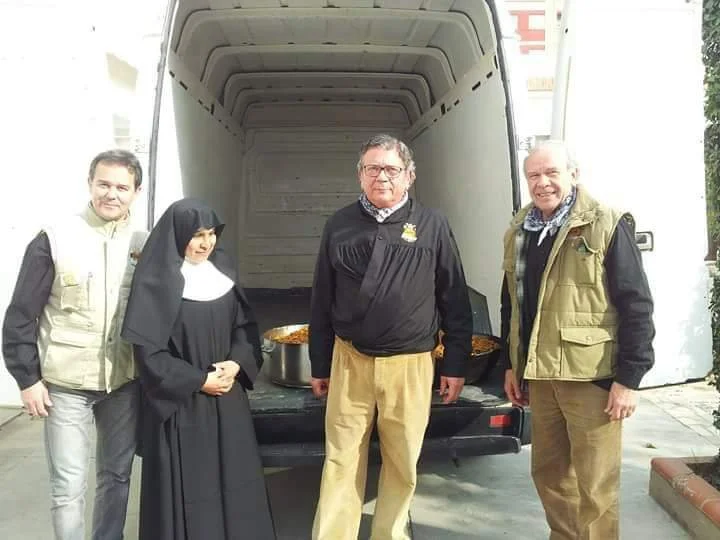 Four people standing in front of an open delivery truck, one dressed as a nun, two men wearing casual jackets, and one man in a black shirt and tan pants, with food trays inside the truck.