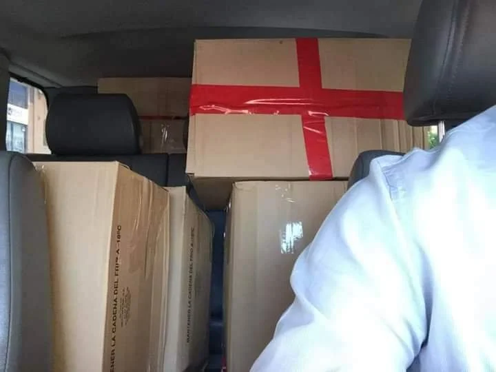 Interior of a vehicle packed with cardboard boxes, with one box sealed with red tape, and the back of a person's shoulder and arm visible in the foreground.