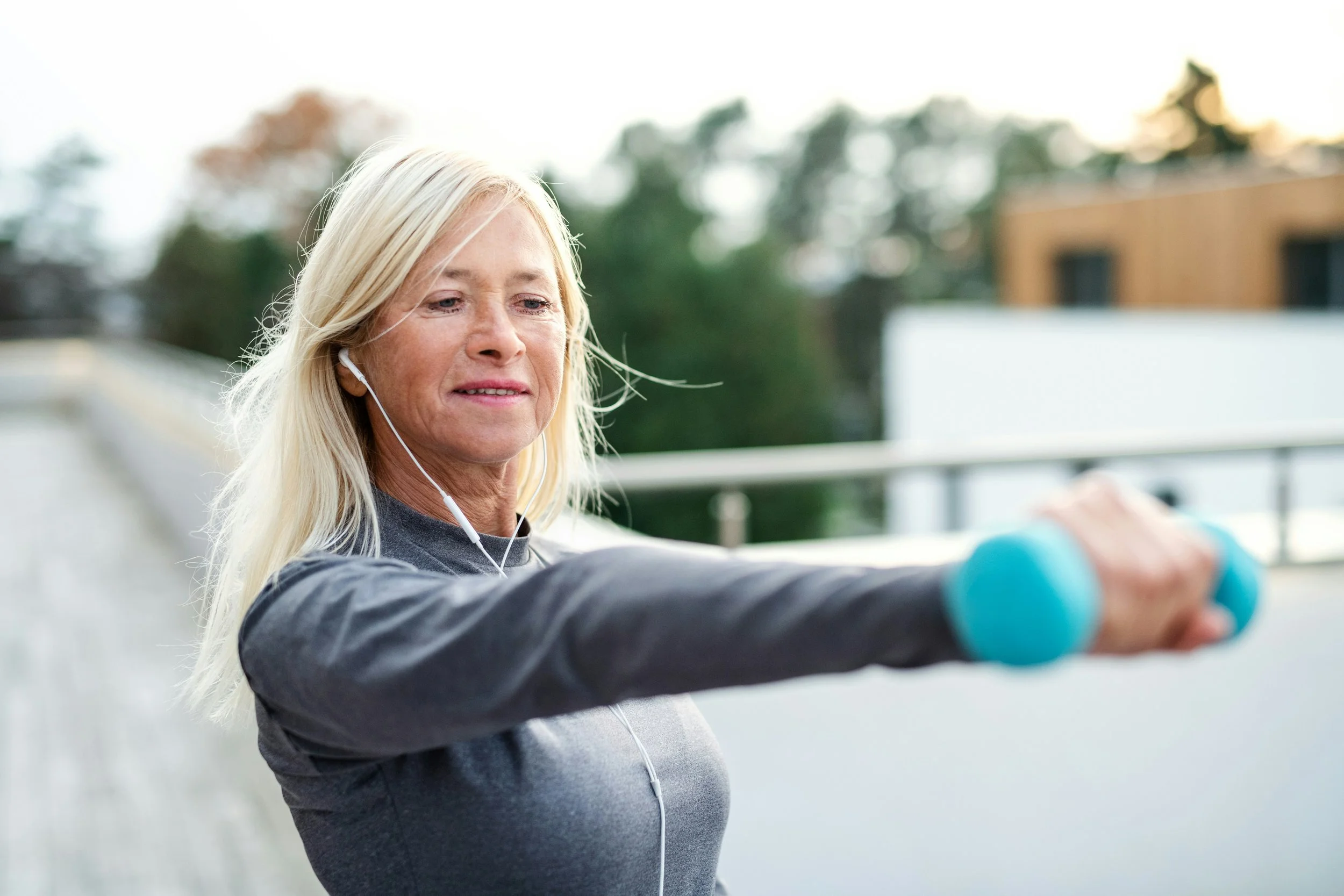 Older woman with long blonde hair, wearing a gray athletic top and earbuds, exercising outdoors with a blue dumbbell, standing on a balcony or terrace.