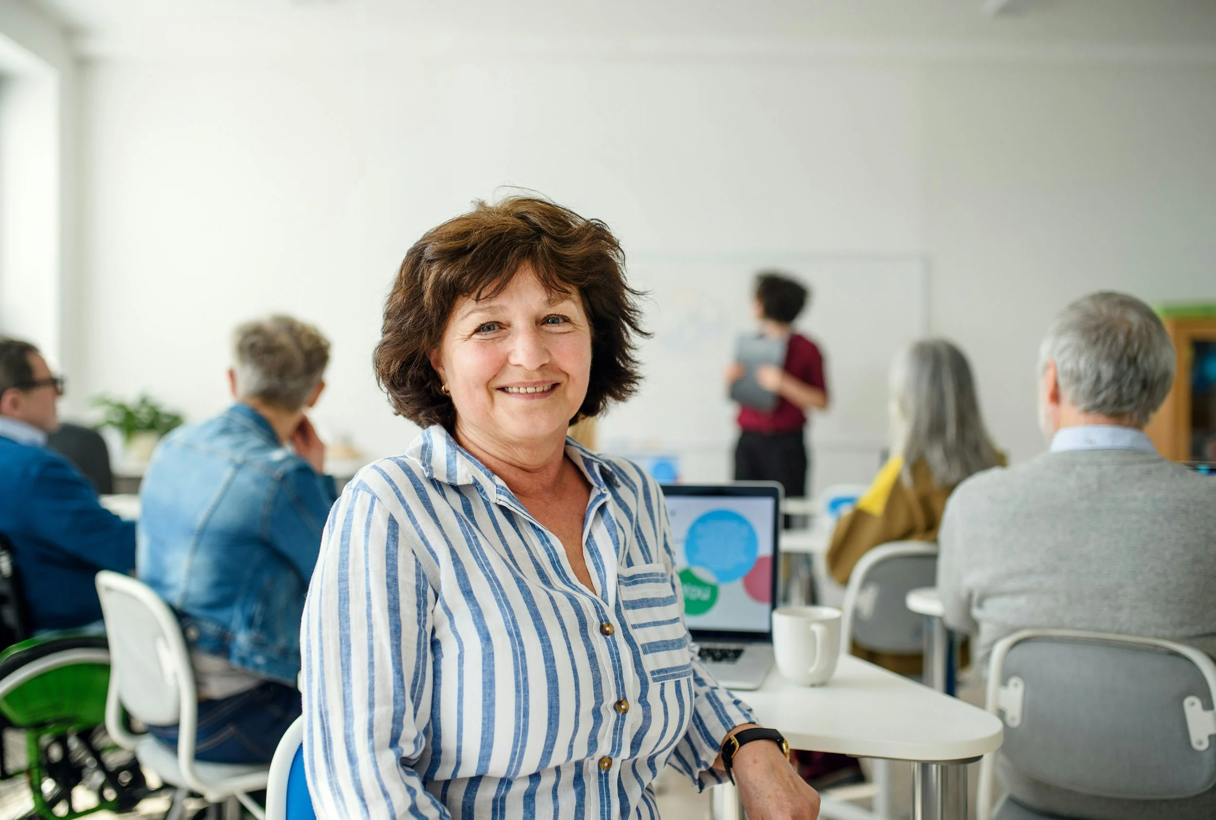 Smiling woman in a striped shirt sitting at a table in a classroom or conference room with other people and a presenter in the background.