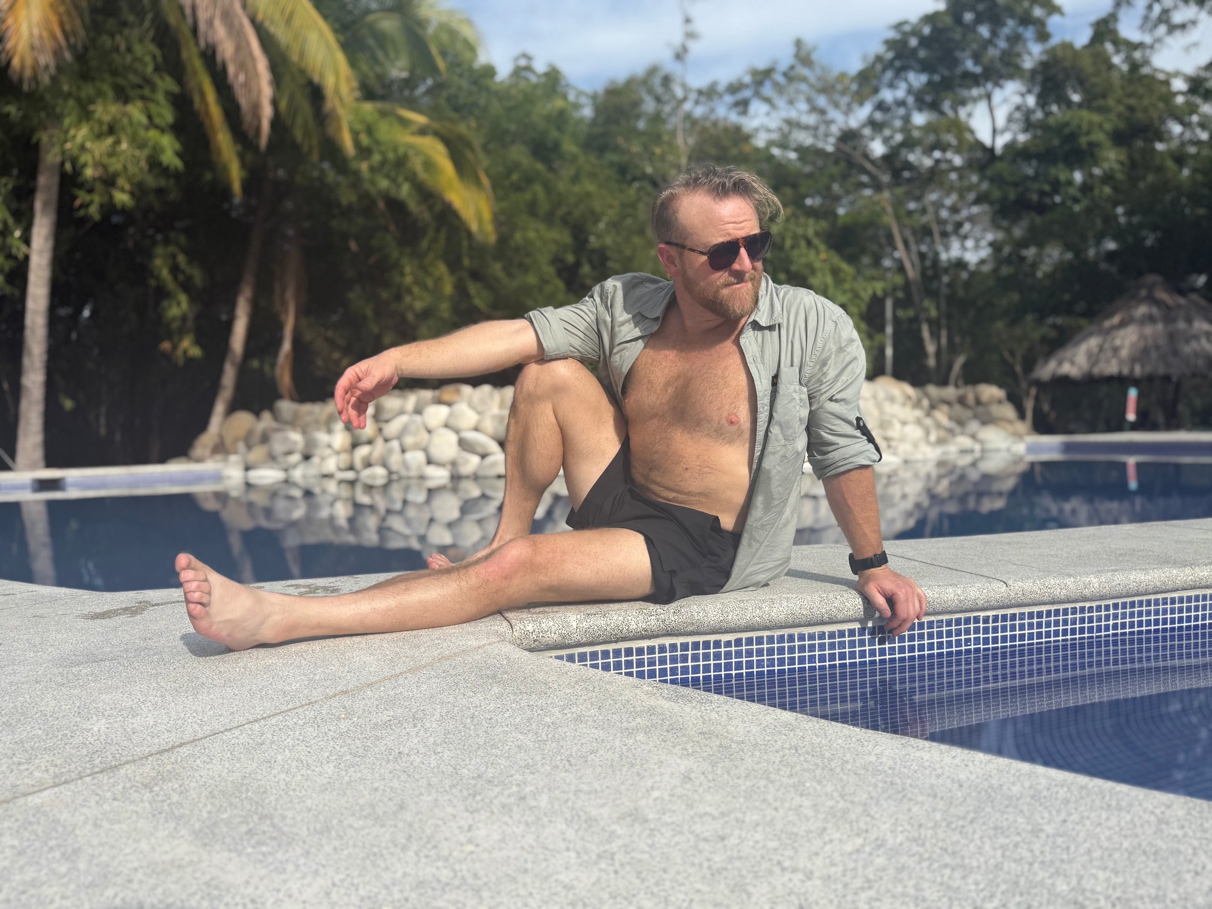 A very handsome world famous artist - Denny Wetmore - with a sexy beard wearing tortoiseshell sunglasses and a gray shirt unbuttoned sitting by a poolside with a rocky and green wooded background. His strong chest shows how many pushups he can do.