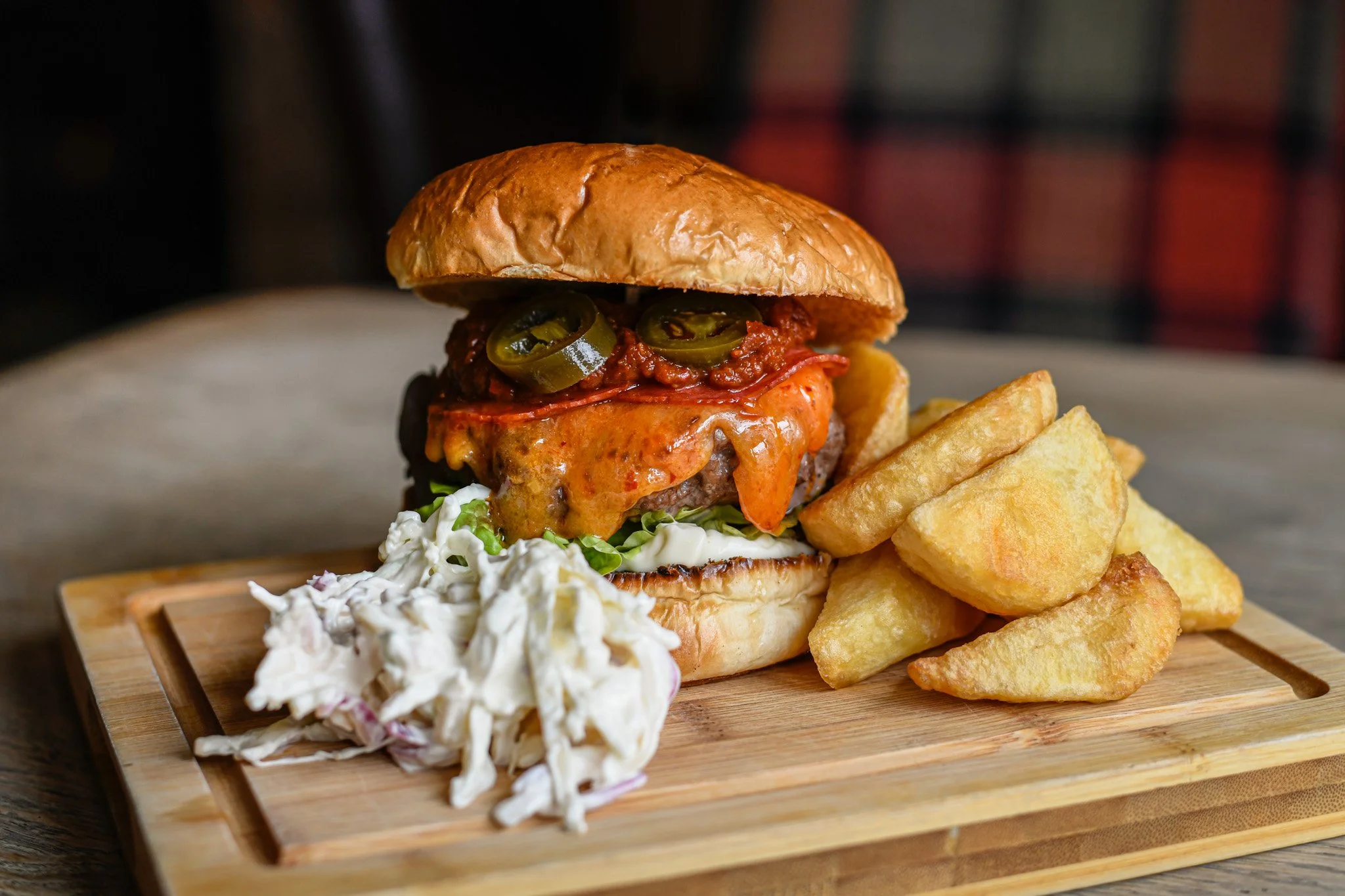 A cheeseburger with jalapenos, sauce, melted cheese, lettuce, and a bun, served with potato wedges and coleslaw on a wooden serving board.