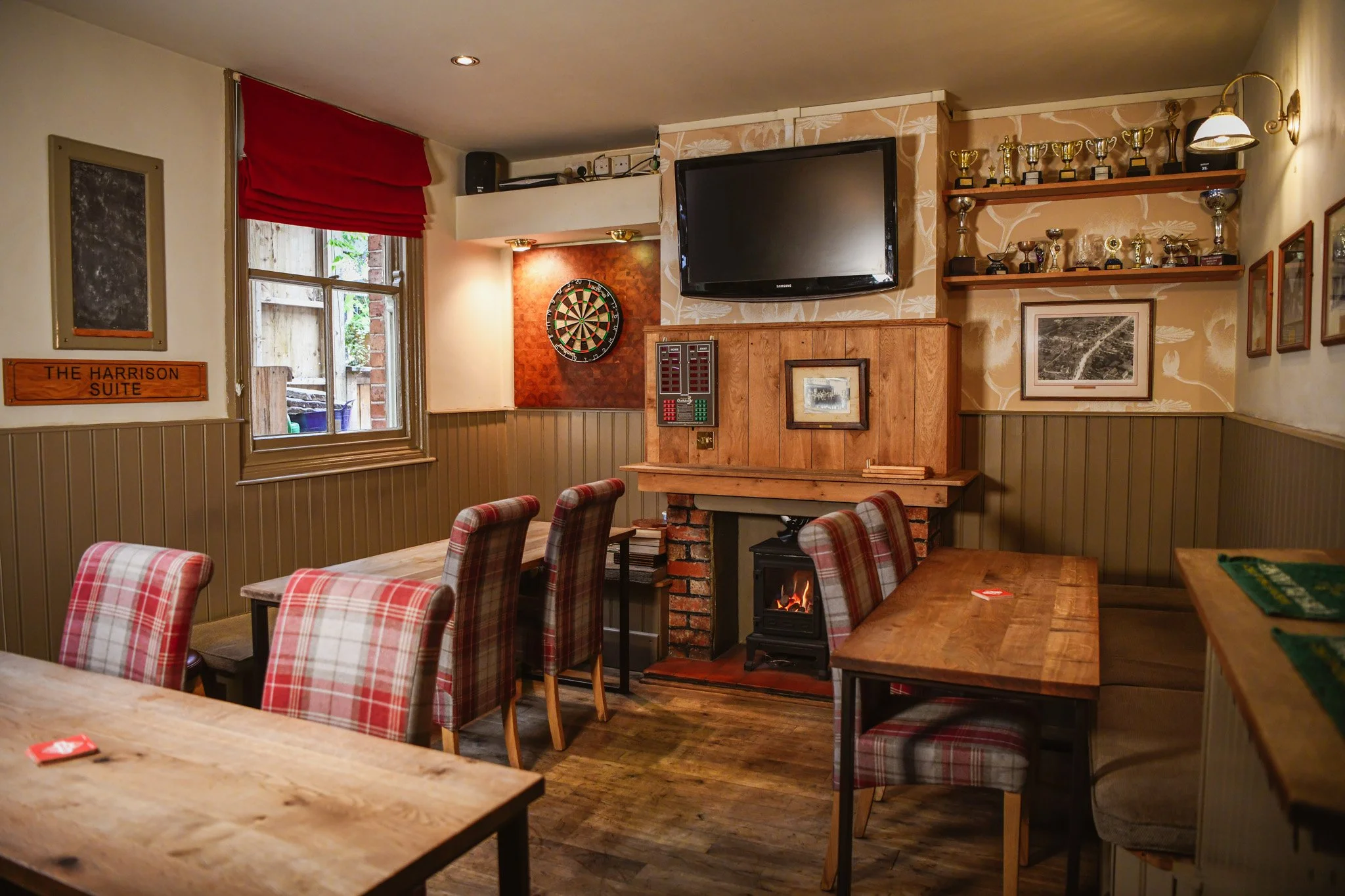 Cozy dining area with plaid upholstered chairs, a wooden table, a fireplace, a dartboard, a TV, and shelves with trophies, in a room with wood paneling and vintage decor.