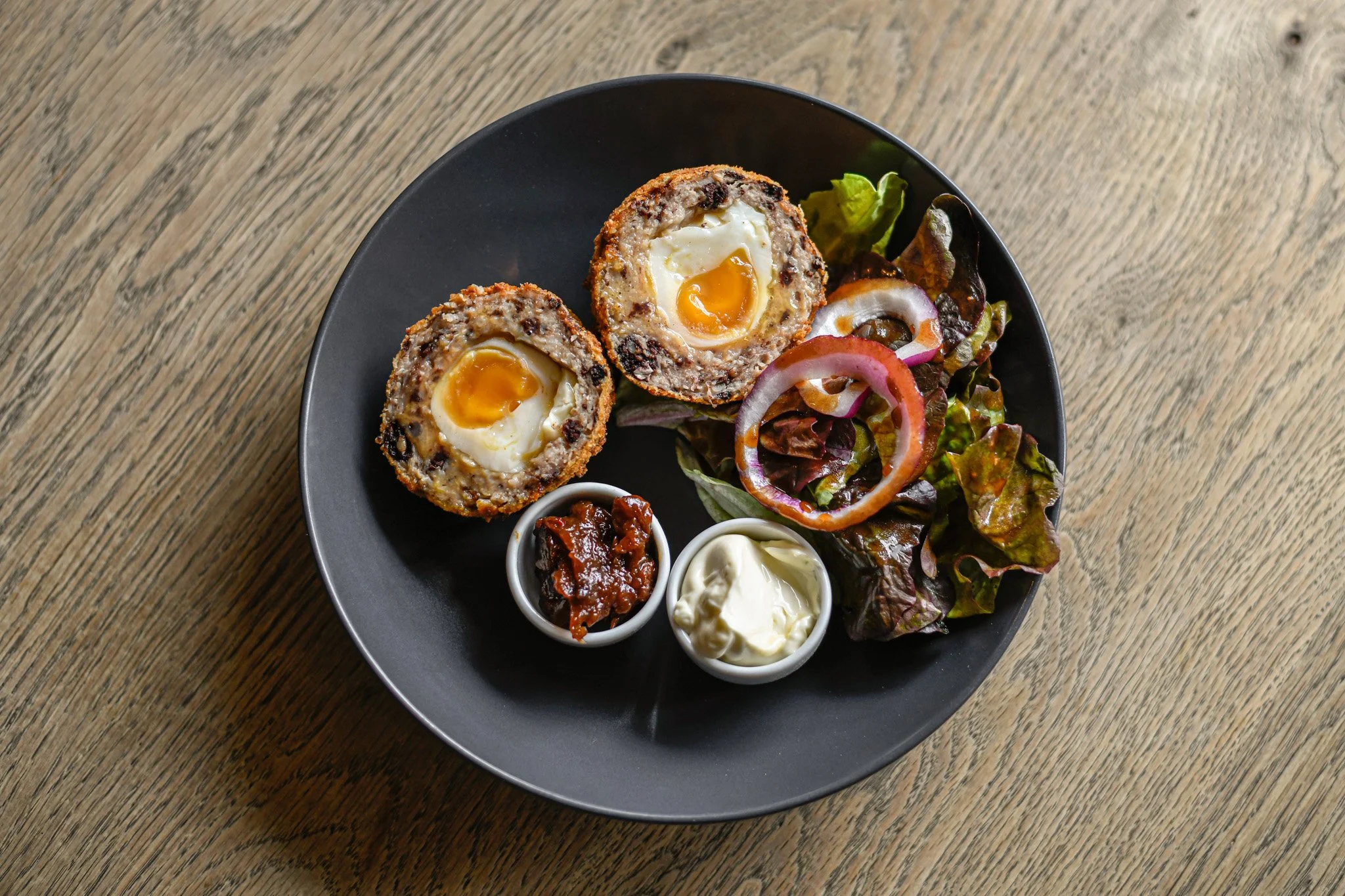 Baked egg stuffed in a meatball, served with a side salad and small bowls of relish and mayonnaise on a black plate on a wooden table.