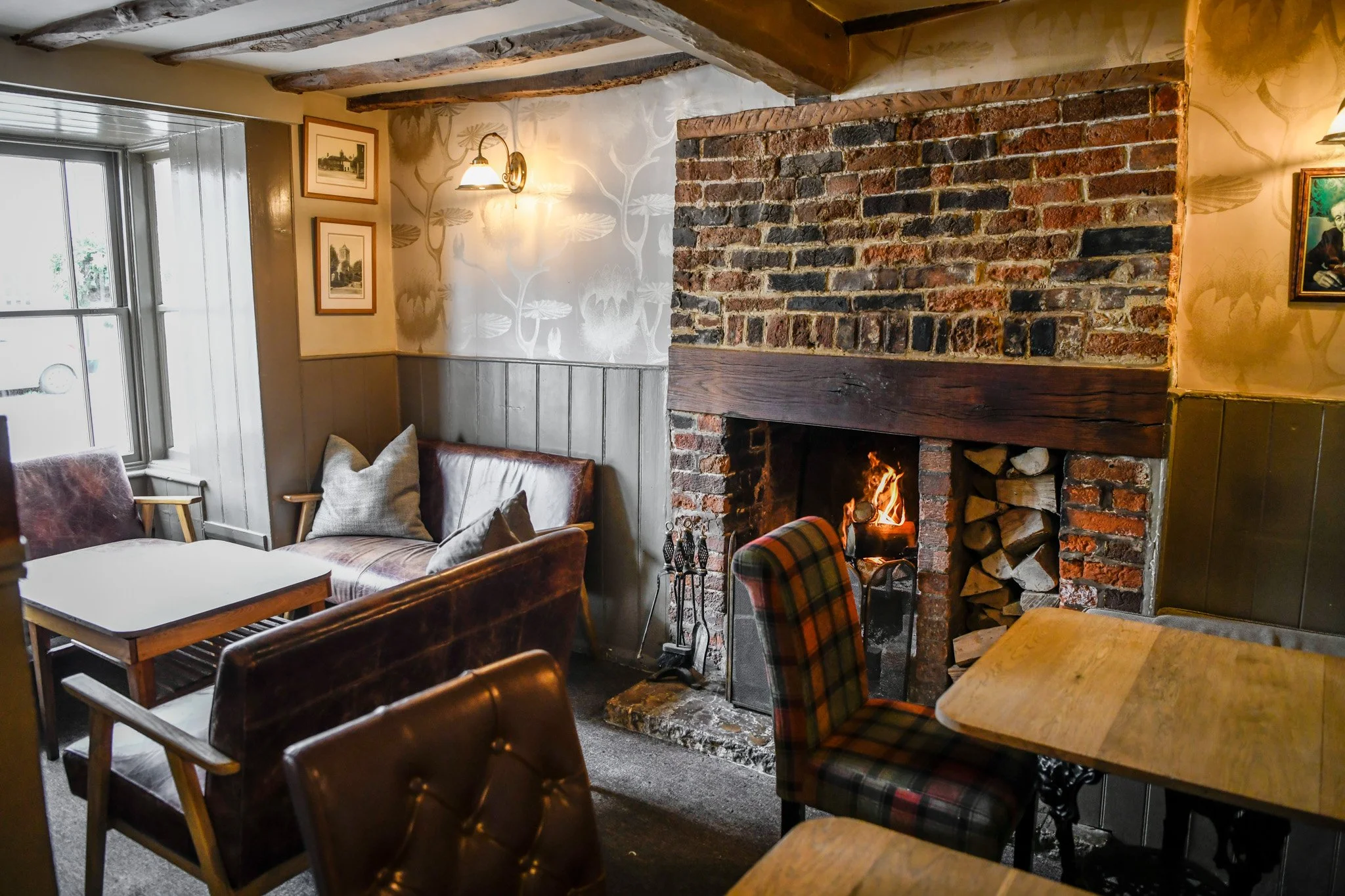 Cozy living room with exposed brick fireplace, wood logs stored on the right, a fire burning inside, wooden and upholstered chairs, a window to the left, and framed artwork on the walls.