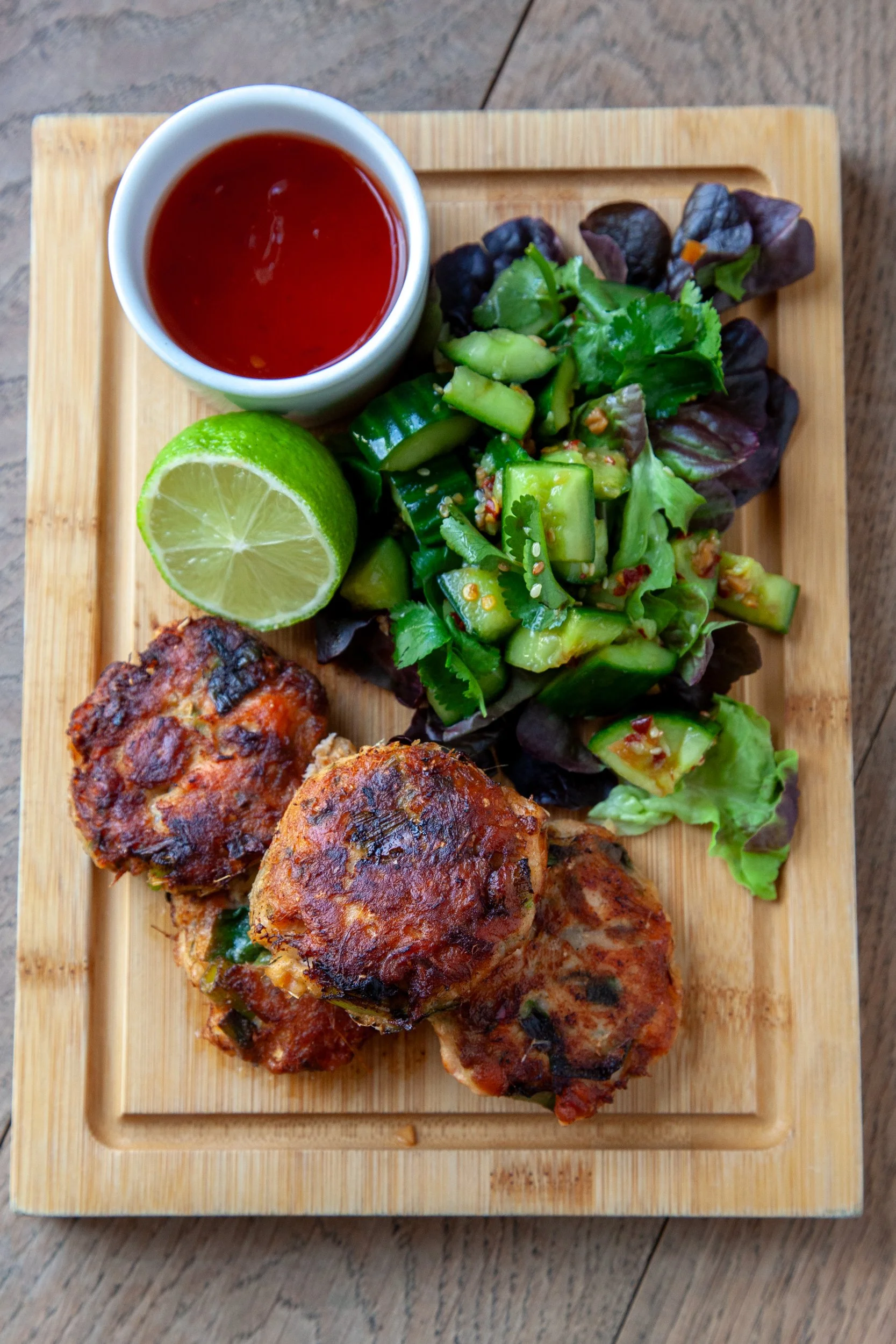 A wooden serving board with grilled chicken meatballs, chopped cucumber salad, lime wedge, and a small cup of red dipping sauce.