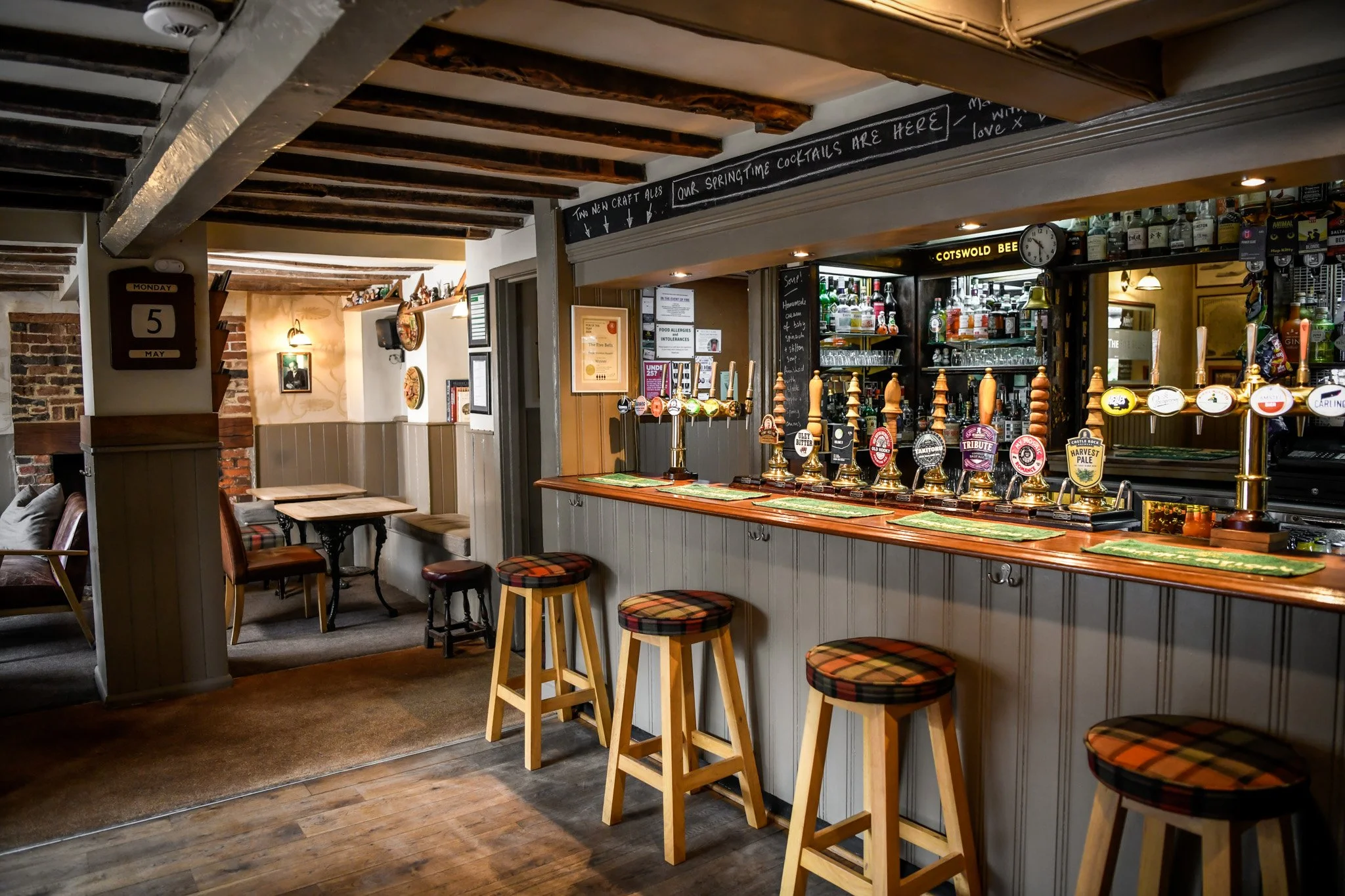 Interior of a pub with a row of bar stools at a wooden bar counter, behind which are many beer pumps and bottles of alcohol. To the left, there are tables and chairs, with a cozy ambiance created by warm lighting and exposed brick walls.