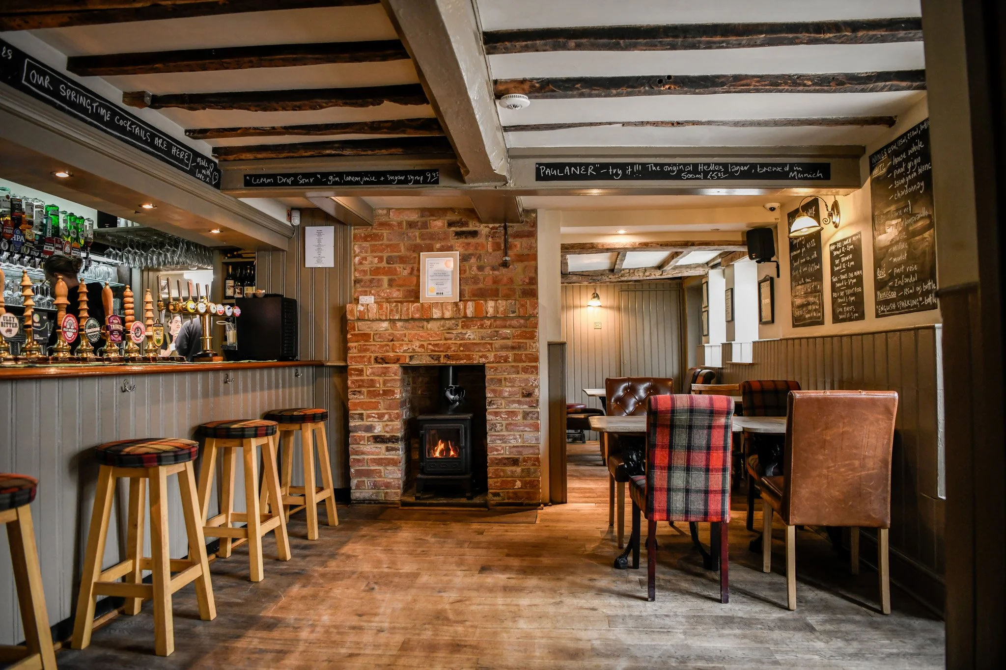 Interior of a cozy pub with a bar area featuring beer taps, a brick fireplace, and a seating area with tables and chairs.
