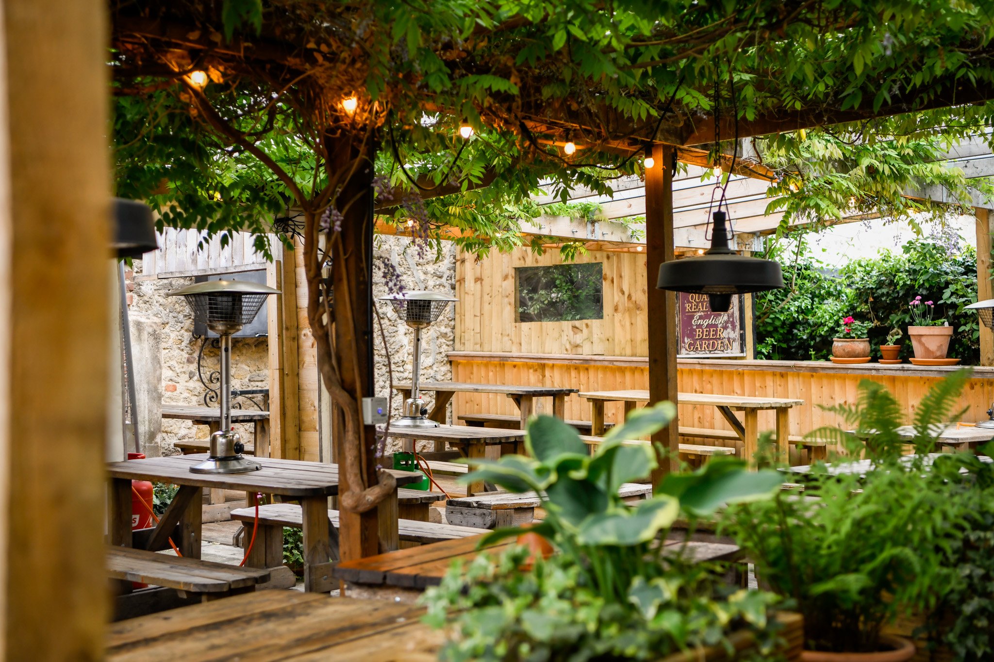 Outdoor patio space with wooden tables, potted plants, string lights, and patio heaters, surrounded by greenery and a wooden fence.