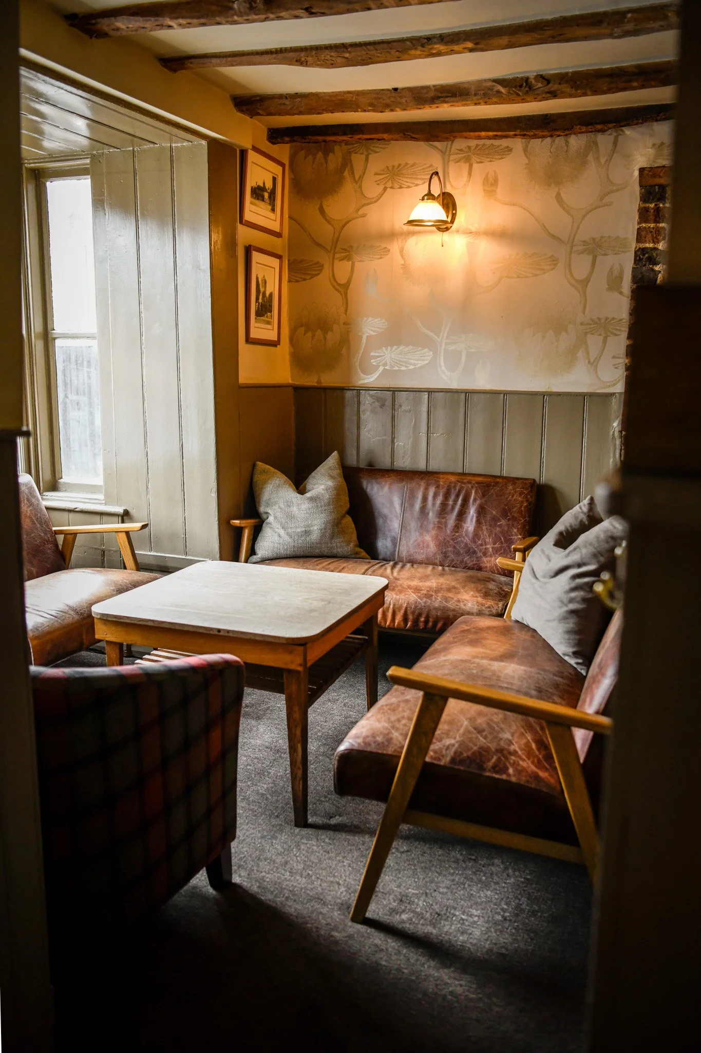 Cozy seating area with a square table, leather and fabric armchairs and a sofa, cushions, wood beams on ceiling, framed pictures on wall, wall lamp, and textured wallpaper.
