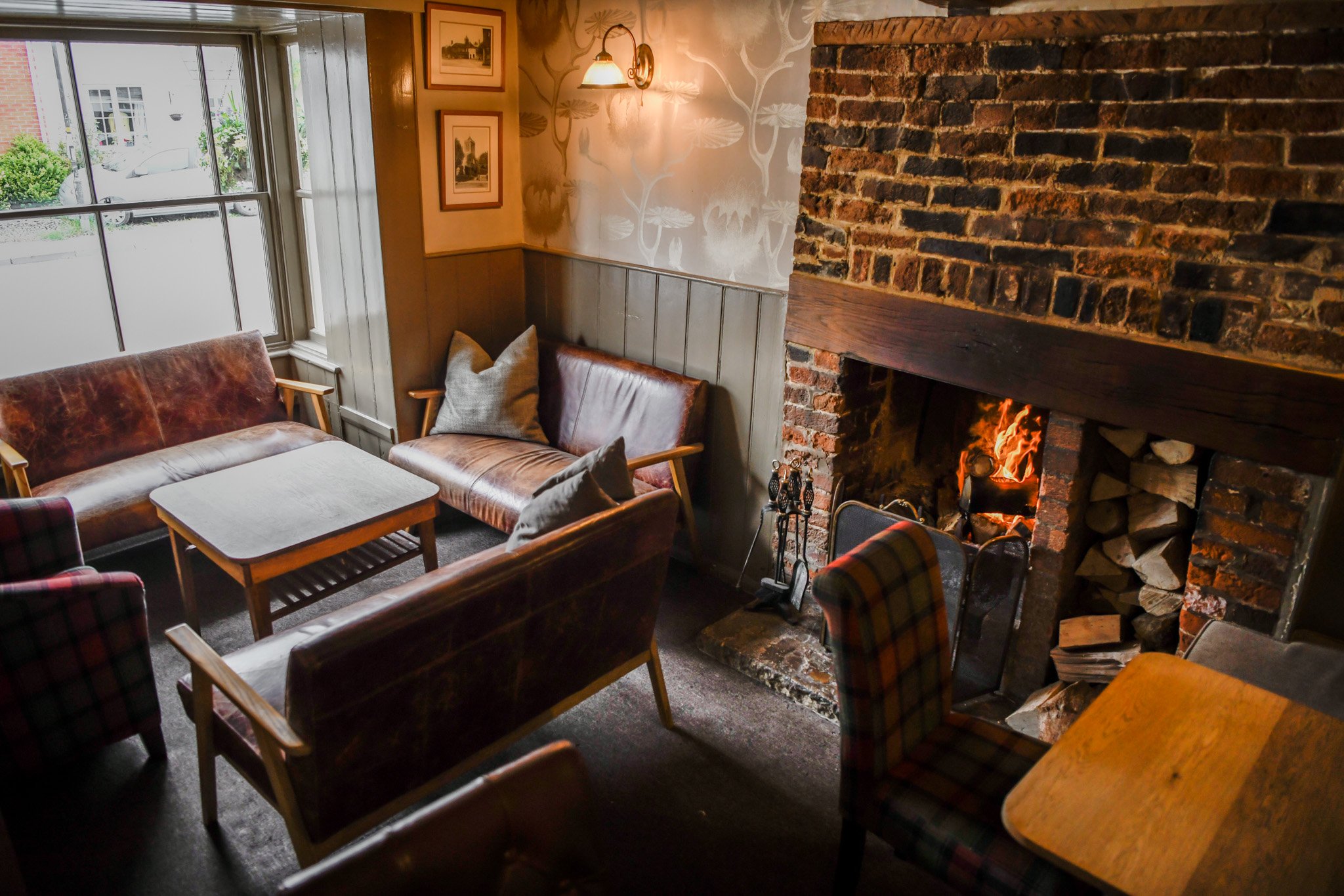 Cozy living room with leather and plaid armchairs, wooden tables, and a brick fireplace with a fire burning, on a dark carpeted floor near a window with outside view.