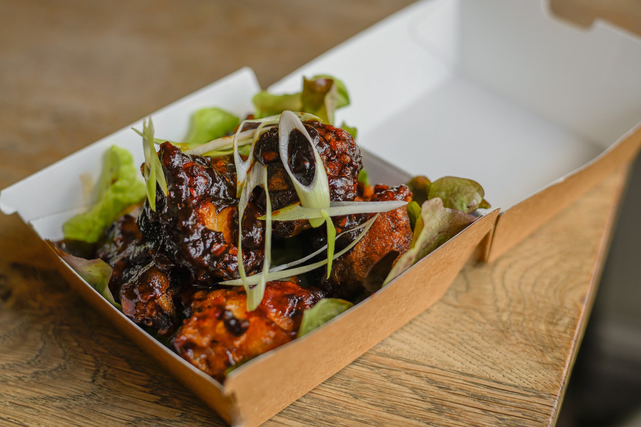 Barbecue chicken wings topped with green onion slices and served on lettuce in a cardboard tray on a wooden table.