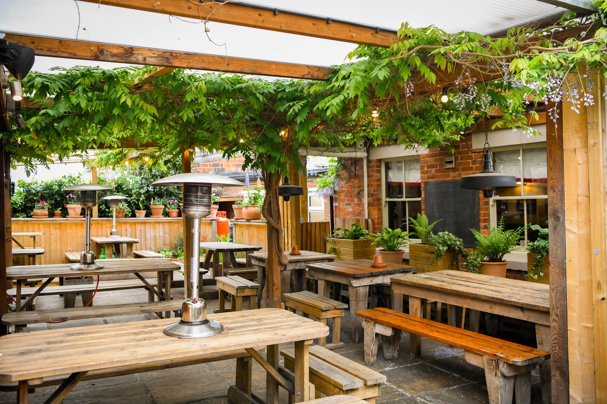 Outdoor patio with wooden tables and benches, potted plants, string lights, and heaters, partially shaded by green foliage and vines.