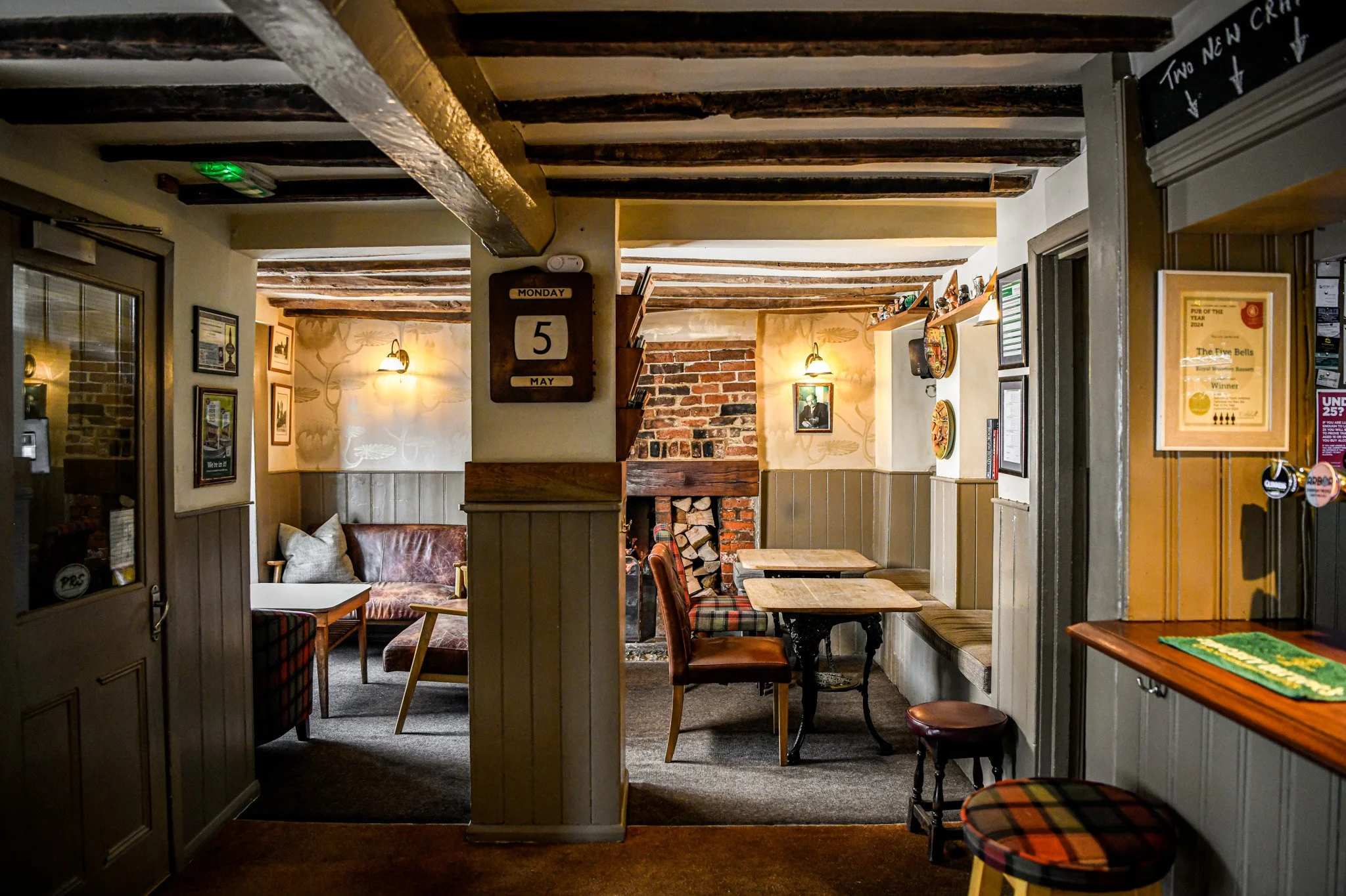 Cozy pub interior with brick fireplace, wooden beams, and mismatched furniture including a plaid armchair and wooden tables.