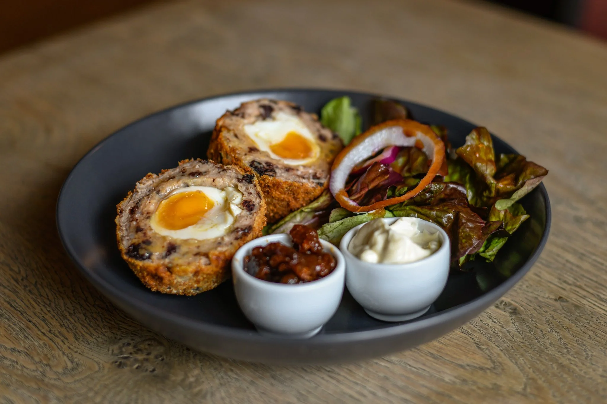 A black plate with two Scotch eggs, side salad with lettuce, tomato, onions, and peppers, and two small bowls of sauces or condiments, all on a wooden table.