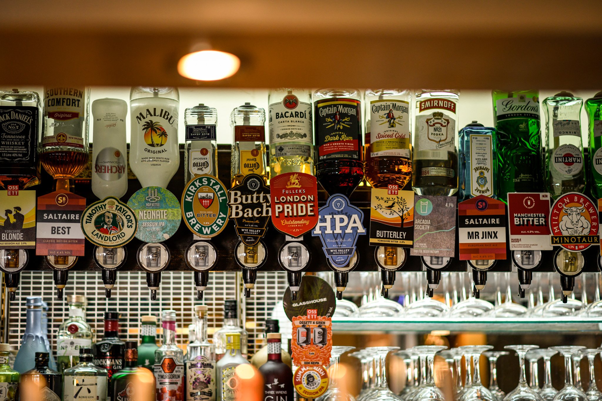 A row of liquor bottles and tap handles with various branding at a bar, including whiskey, vodka, and craft beer.