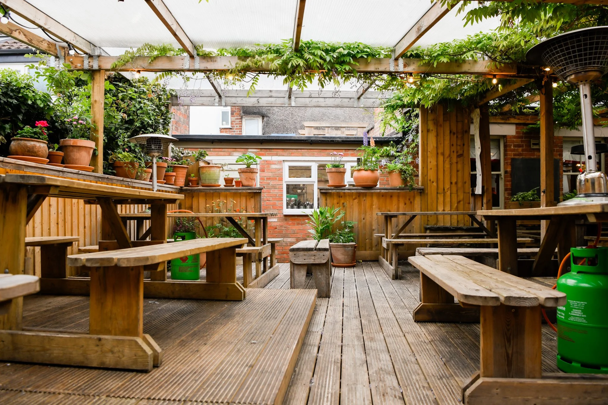 A cozy outdoor wooden patio with picnic-style tables and benches, decorated with potted plants and string lights under a partial transparent roof.
