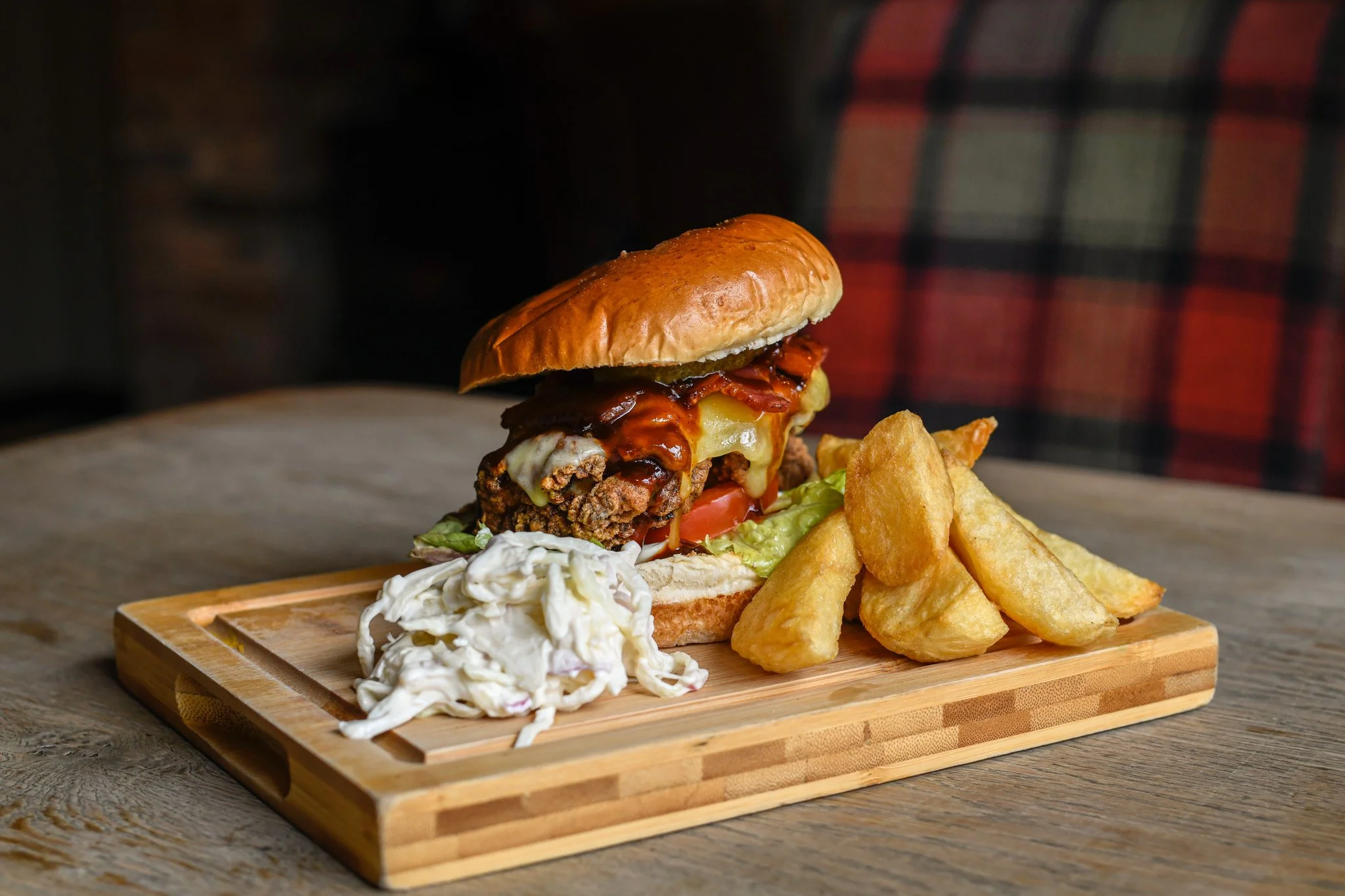 A crispy fried chicken sandwich with bacon, cheese, lettuce, tomato, barbecue sauce on a bun, served with potato wedges and coleslaw on a wooden board.