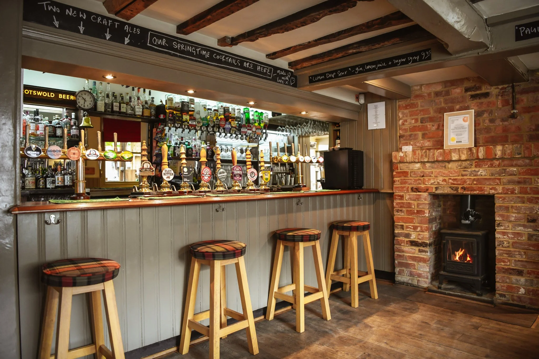 Image of a cozy bar with a row of five wooden stools with plaid cushions at the bar counter, which has multiple beer taps, behind which are shelves of liquor bottles, beer glasses, and hanging wine glasses, with a brick wall and a small fireplace in 