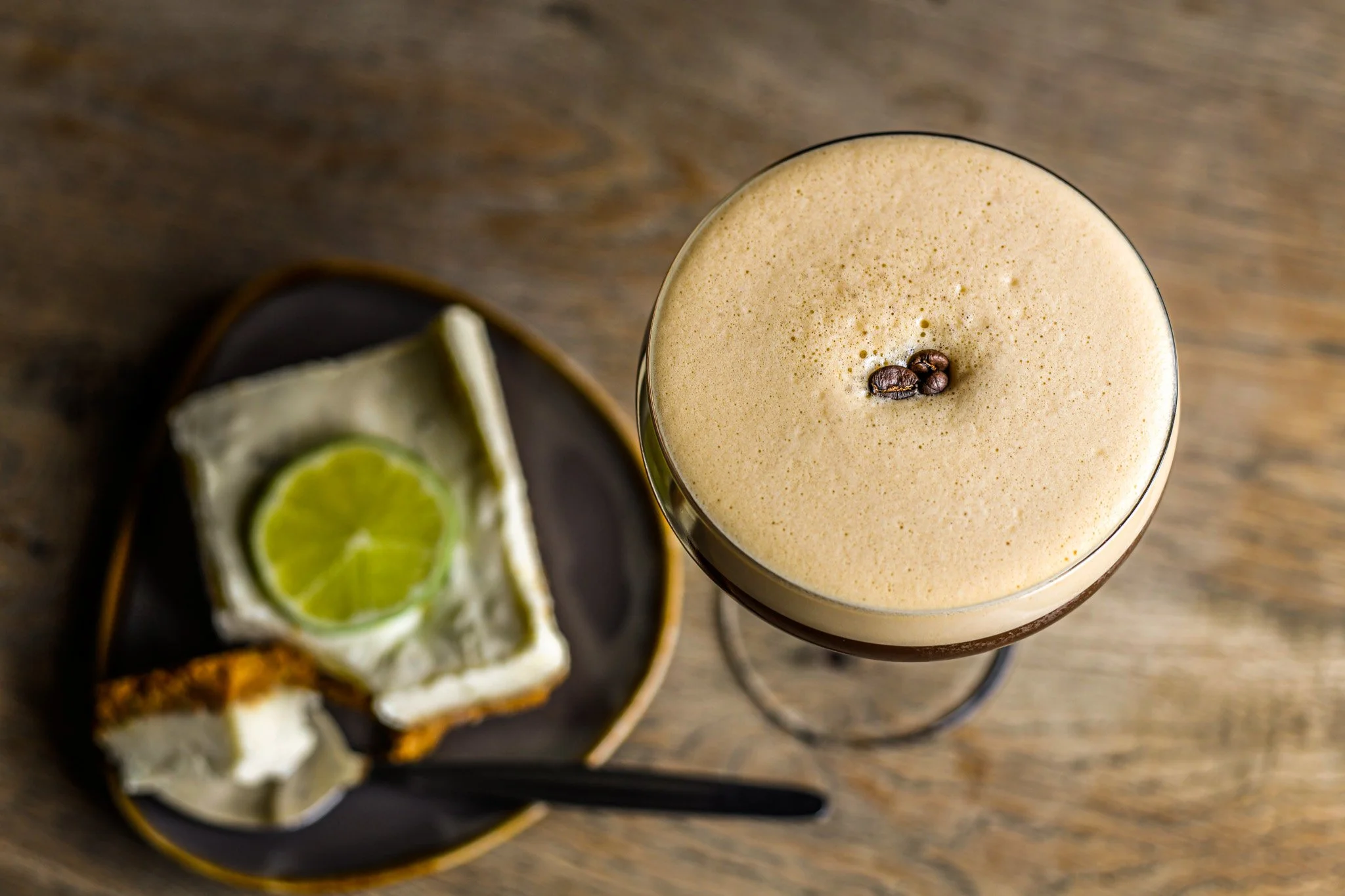 A glass of coffee with foam and three coffee beans on top, next to a plate with a slice of lime and a small piece of cake on a wooden surface.
